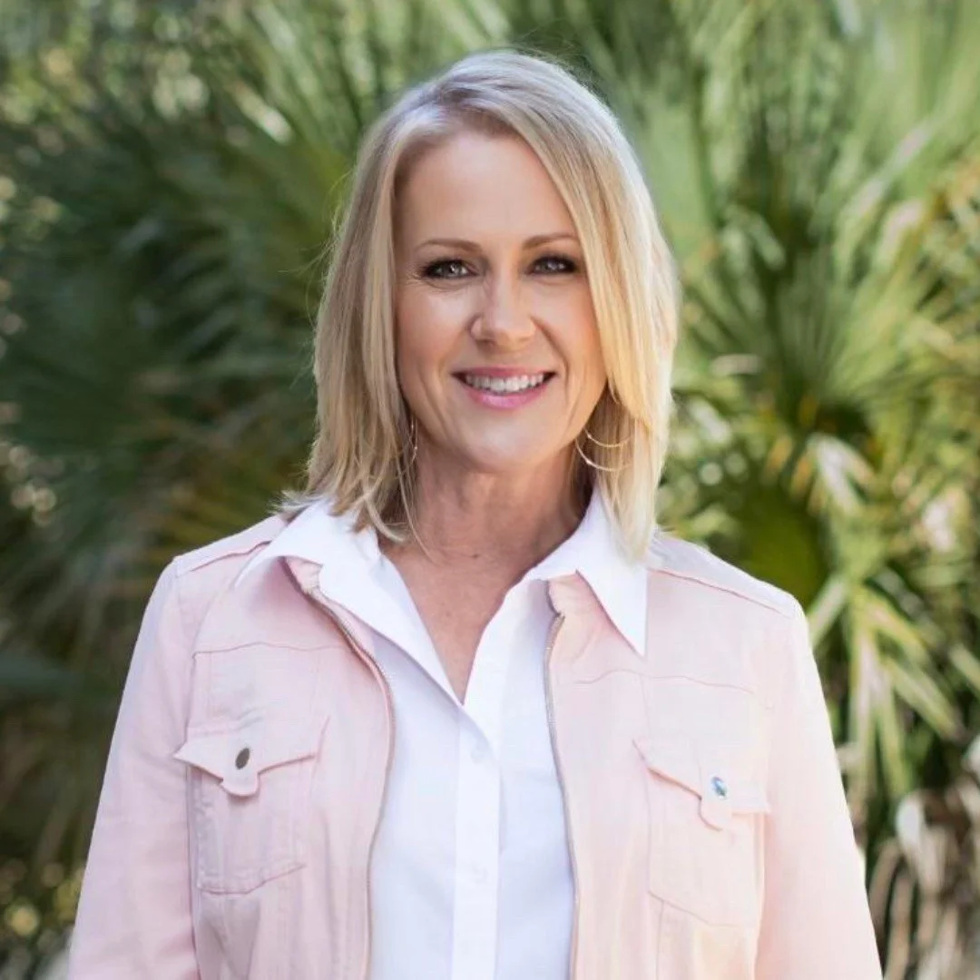 A smiling woman with shoulder-length blonde hair, wearing a light pink jacket and white shirt, outdoors with green foliage in the background.