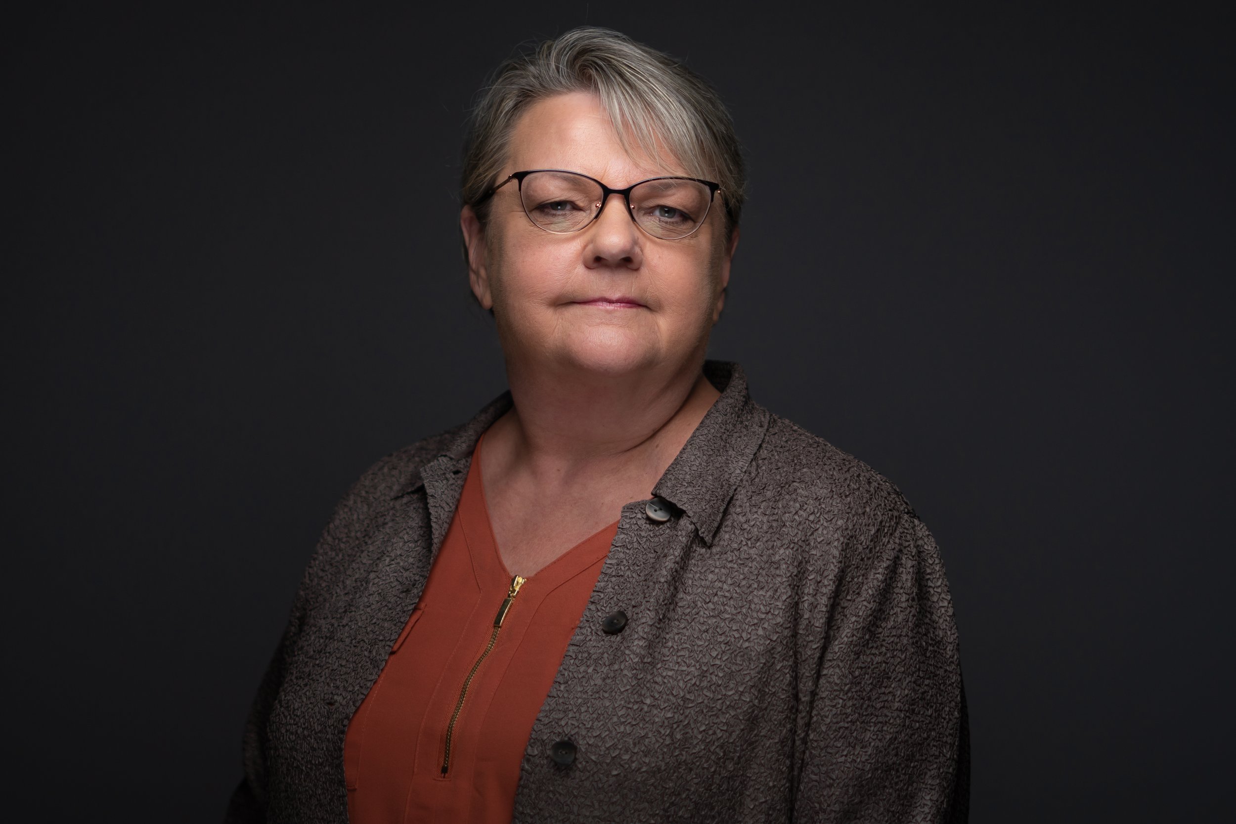 Portrait of an older woman with short gray hair, glasses, and a confident expression, wearing a brown textured jacket over an orange top, against a dark background.