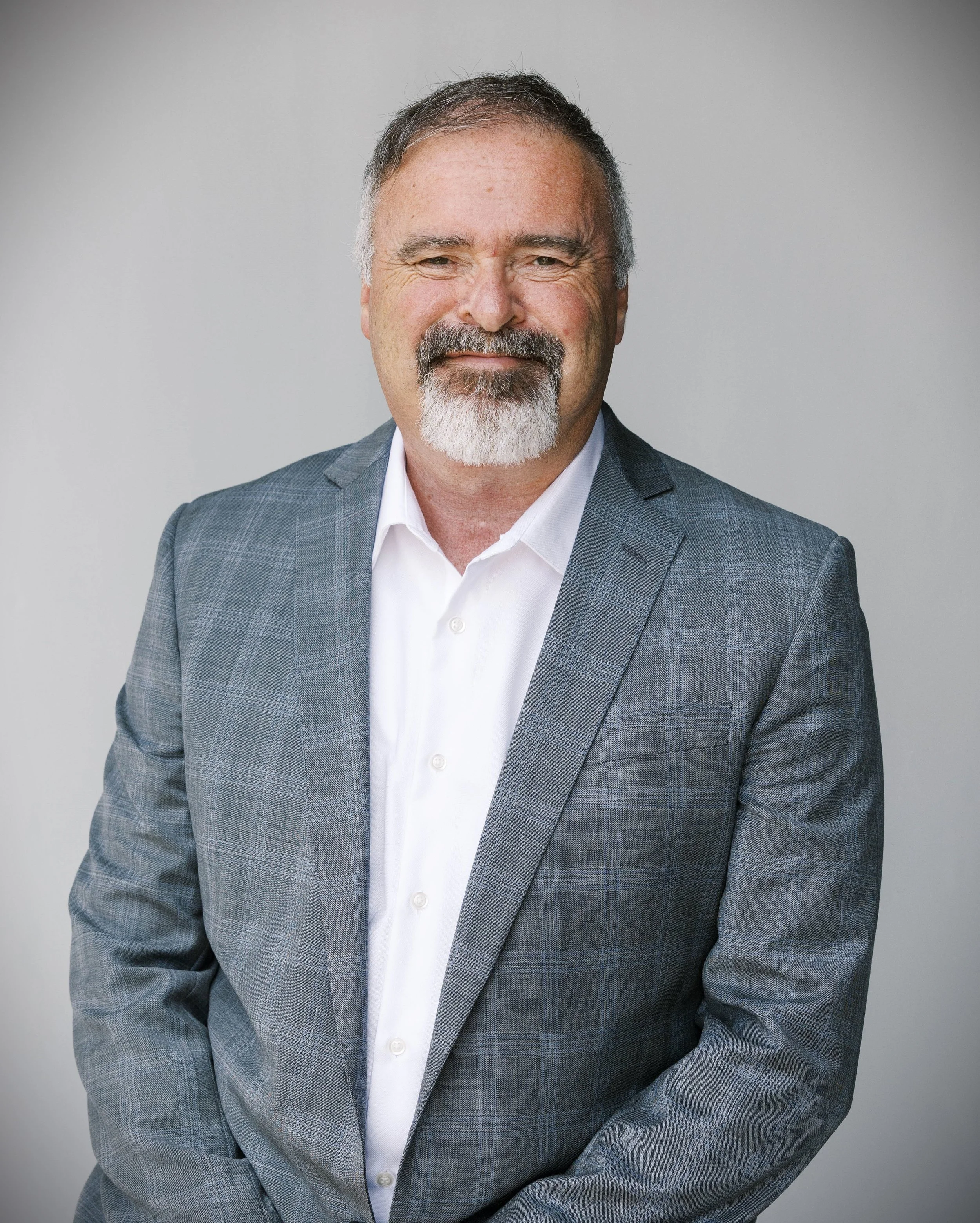 Portrait of a middle-aged man with gray hair and a beard, wearing a gray checkered blazer and white shirt, smiling against a light gray background.