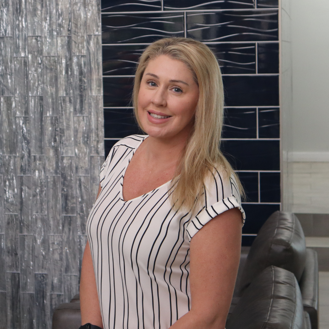 A woman with blonde hair smiling and wearing a white blouse with black vertical stripes, standing in front of a modern wall with blue and gray tile designs.