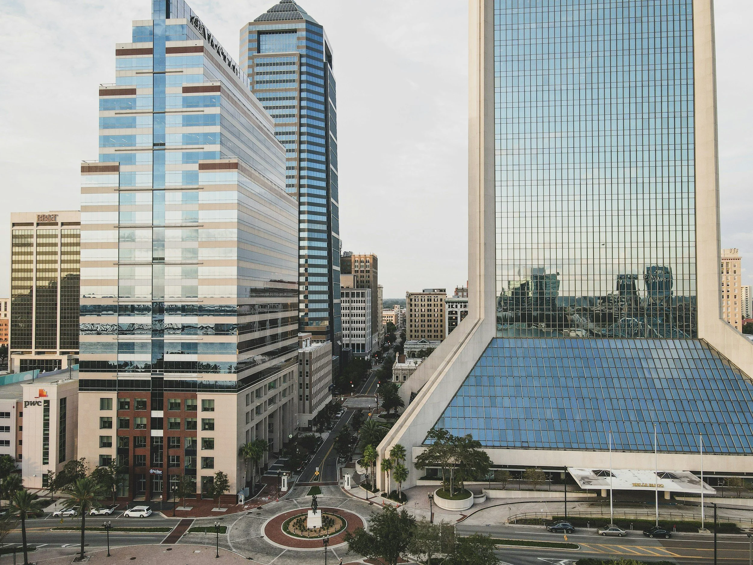 Cityscape of modern skyscrapers with glass facades reflecting the sky, street view with few cars and palm trees, and a small park with a statue in the foreground.