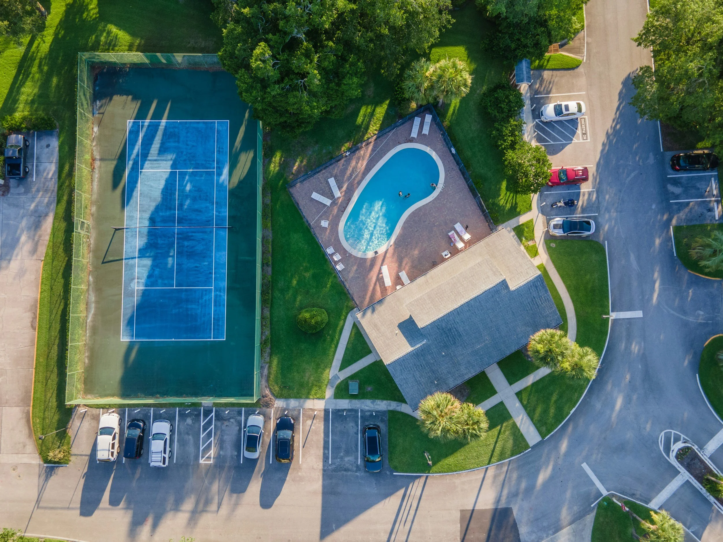 An aerial view of a community park showing a tennis court, a small swimming pool with lounge chairs, a building with a grey roof, a parking lot with several cars, and surrounding trees and greenery.