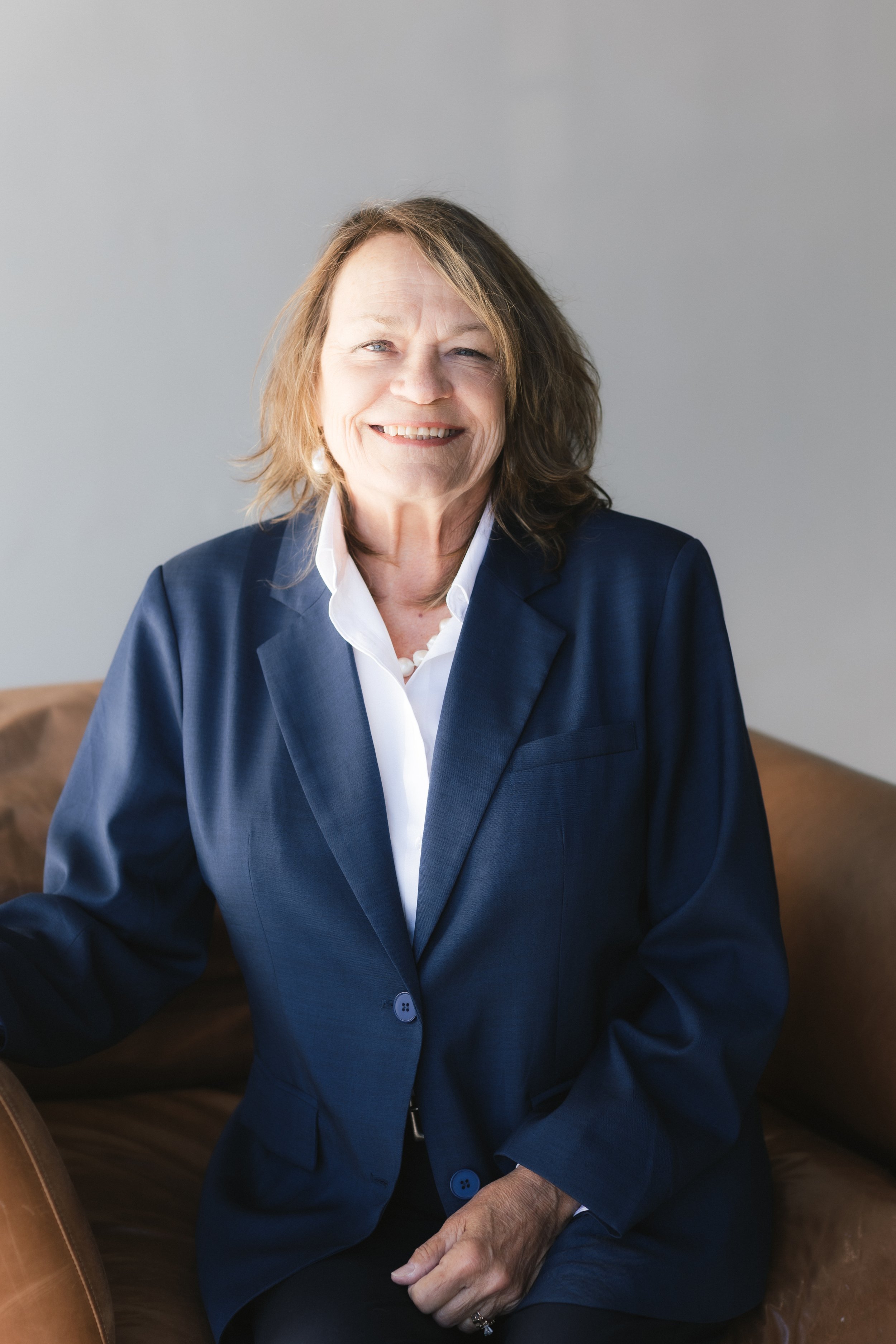 A smiling woman with shoulder-length hair, wearing a navy blue blazer and white shirt, sitting on a brown leather couch against a plain gray background.
