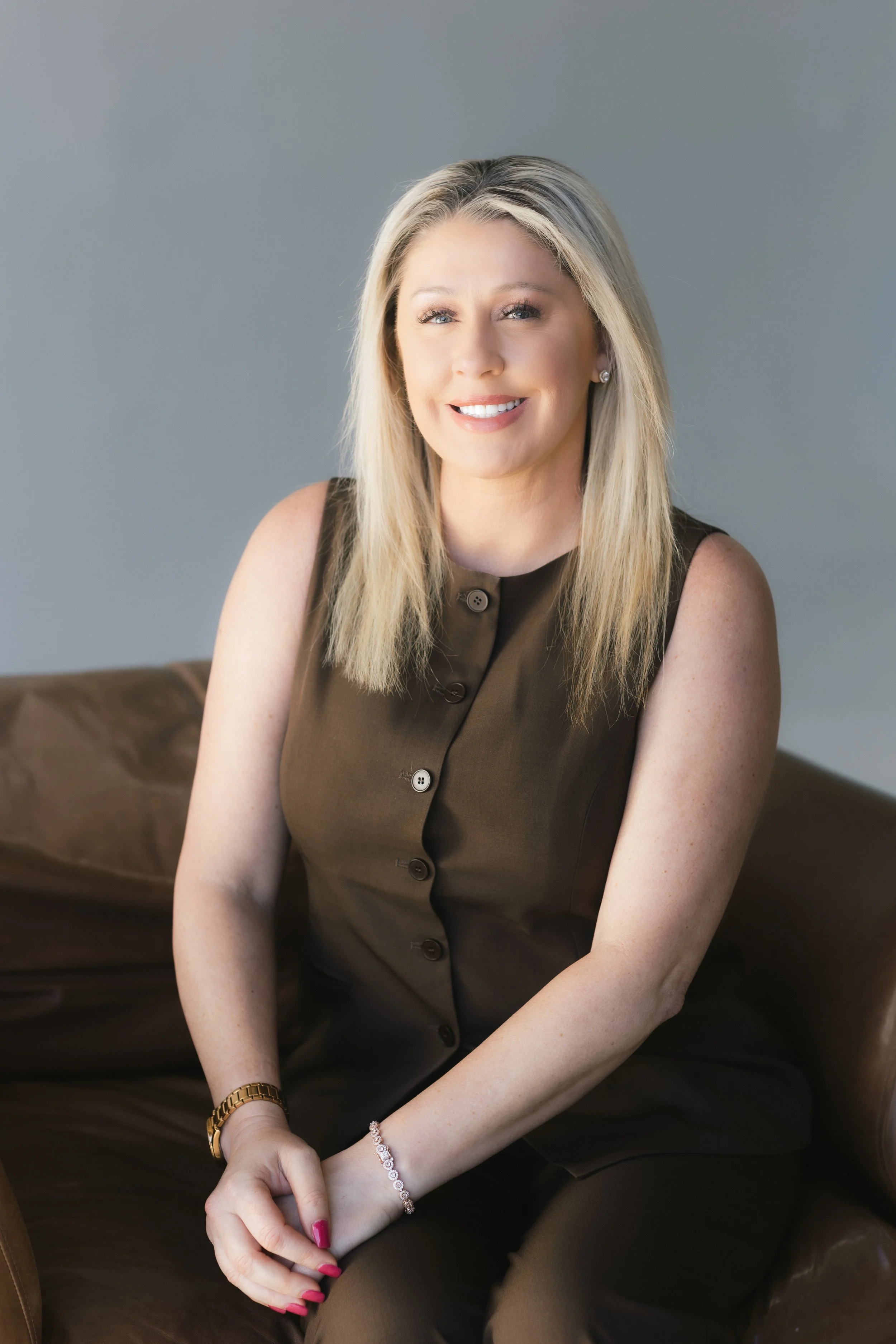 A woman with blonde hair, wearing a sleeveless dark brown top, sitting on a brown leather sofa, smiling at the camera.