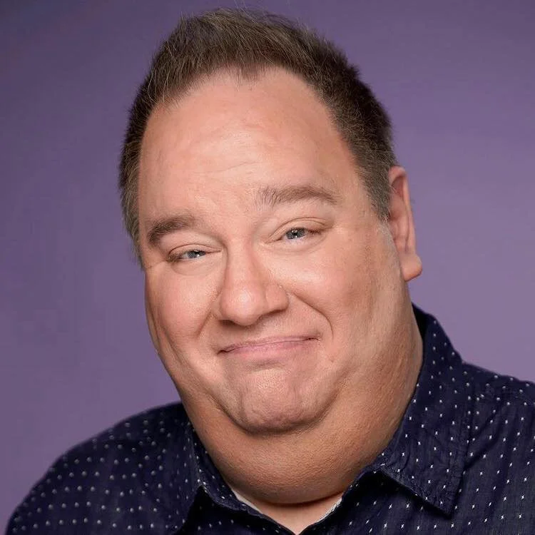 A man with short brown hair, fair skin, and wearing a dark blue shirt with small white dots, smiling at the camera against a purple background.