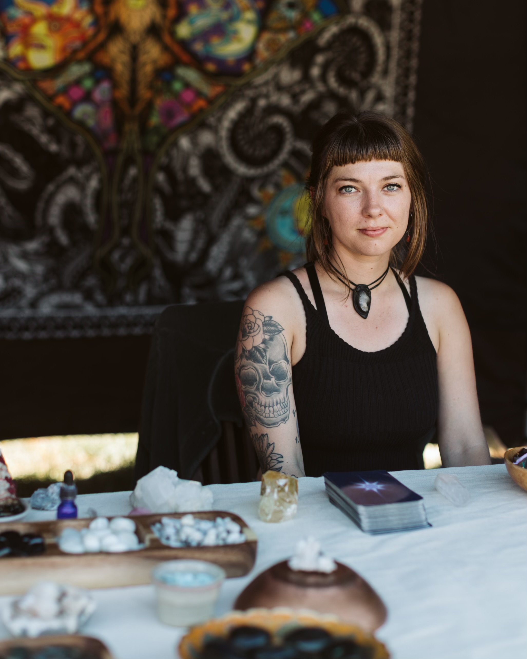 A woman with tattoos on her arm and wearing a black tank top, sitting at a table with tarot cards and crystals, with a colorful, intricate tapestry in the background.
