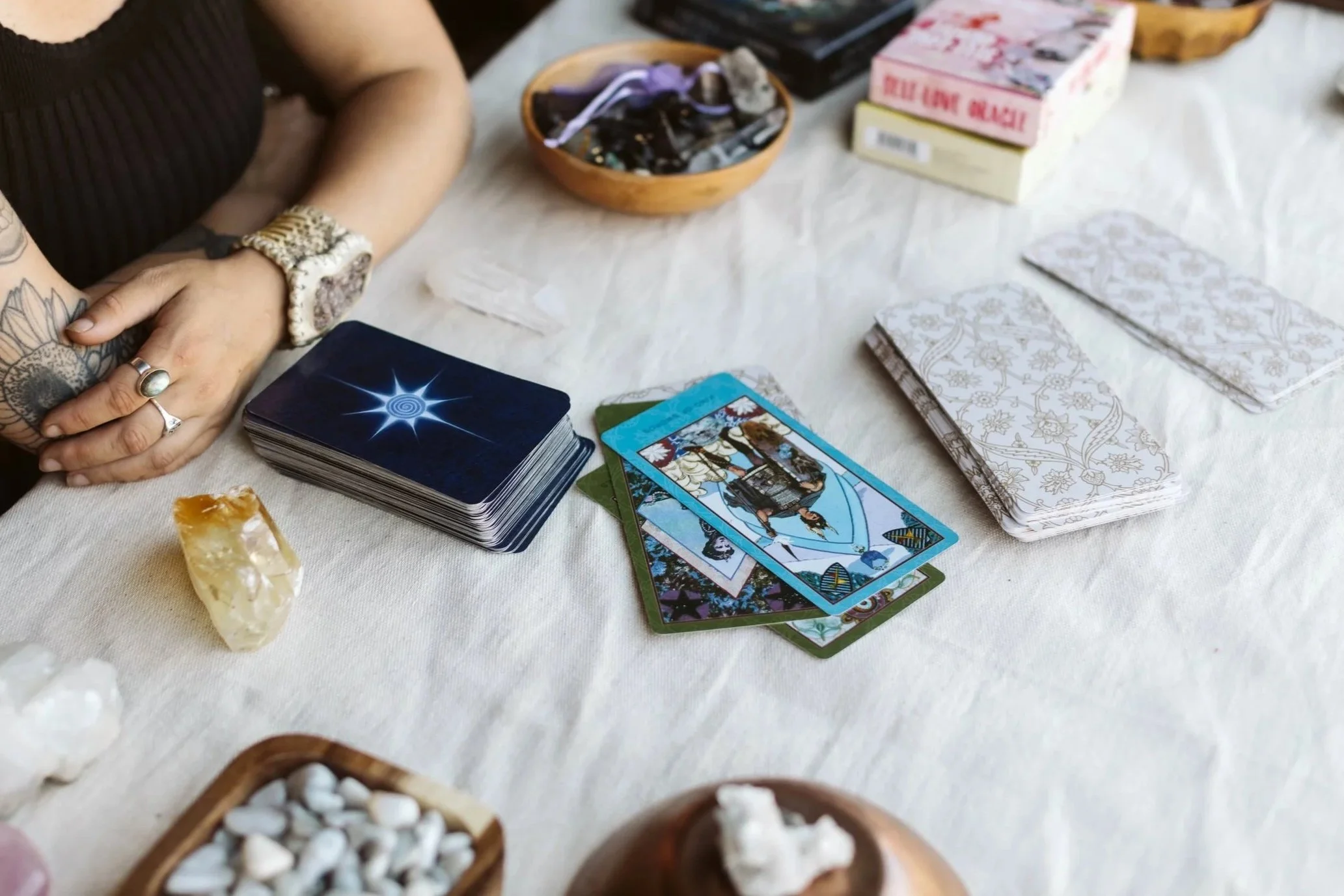 A person with tattoos on their arm and wearing rings and a watch, sitting at a table with tarot cards, crystals, and small decorative bowls.