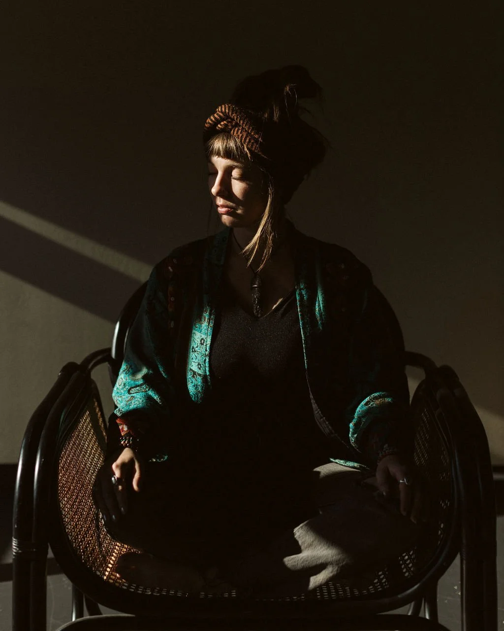 A woman sitting in a chair with her eyes closed, illuminated by sunlight streaming through a window, casting shadows on her face and background.