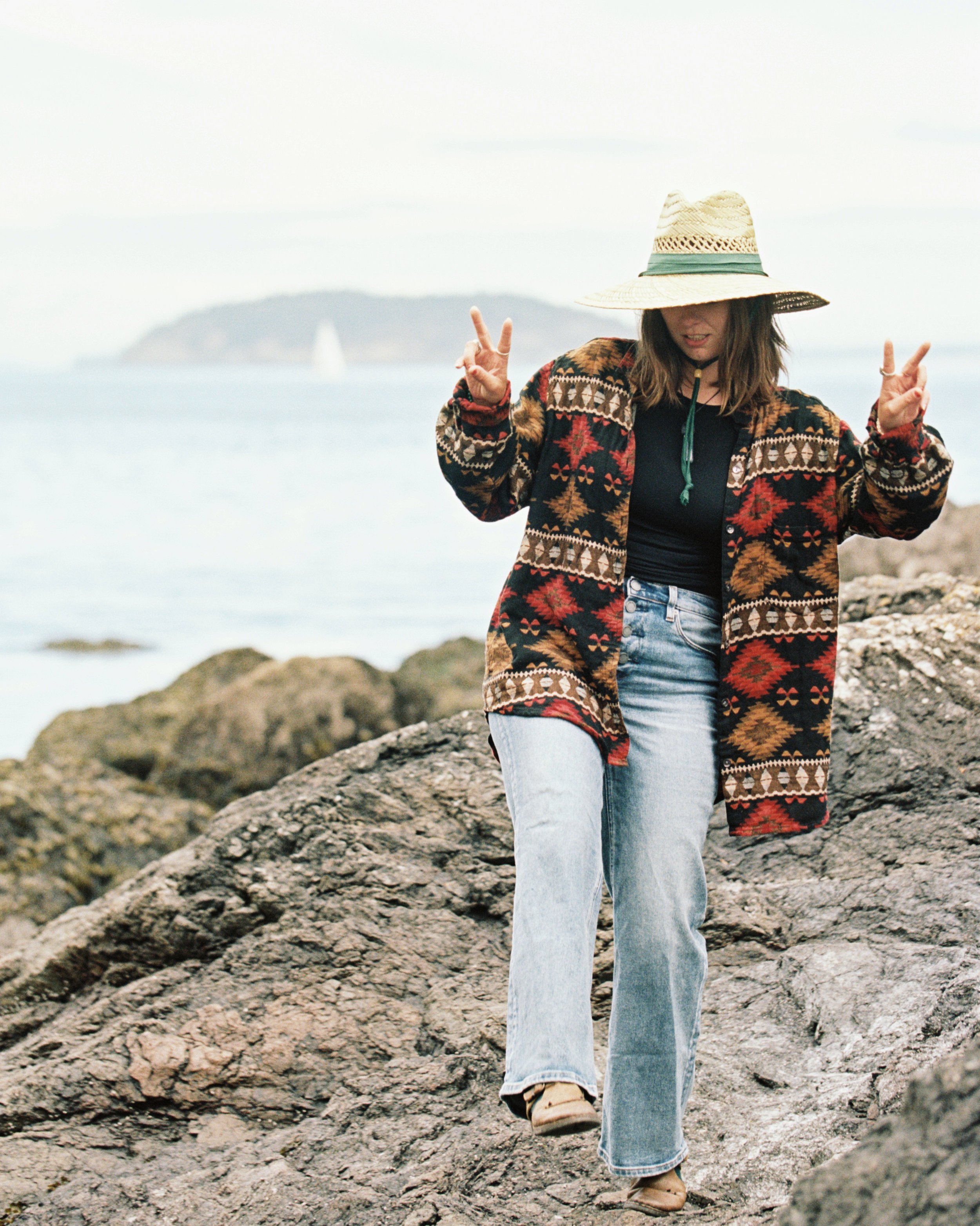 Woman wearing a patterned jacket and a large sun hat making peace signs with both hands on rocky shoreline with ocean in background.