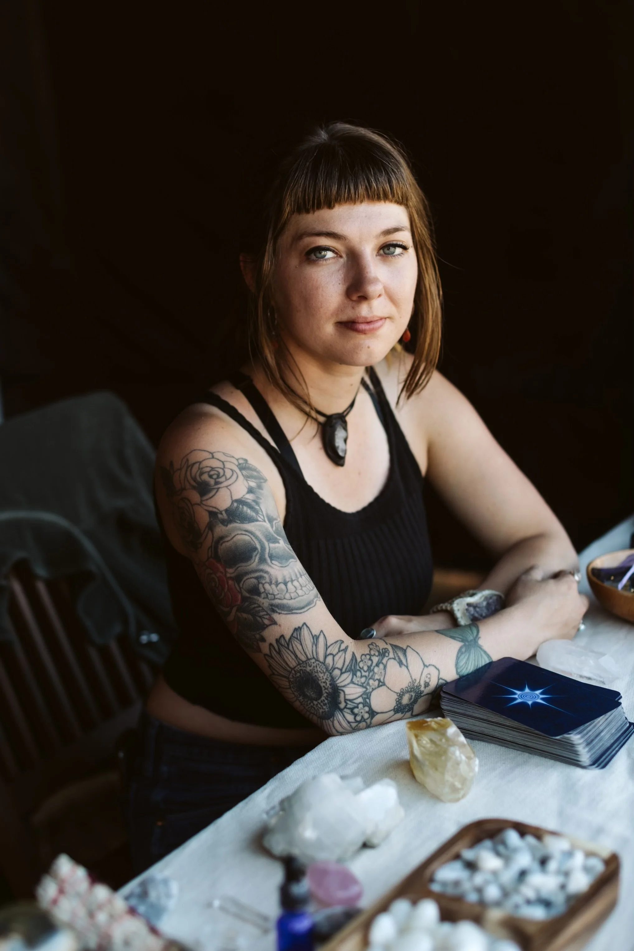 A woman with tattoos on her arm sitting at a table with tarot cards and crystals.