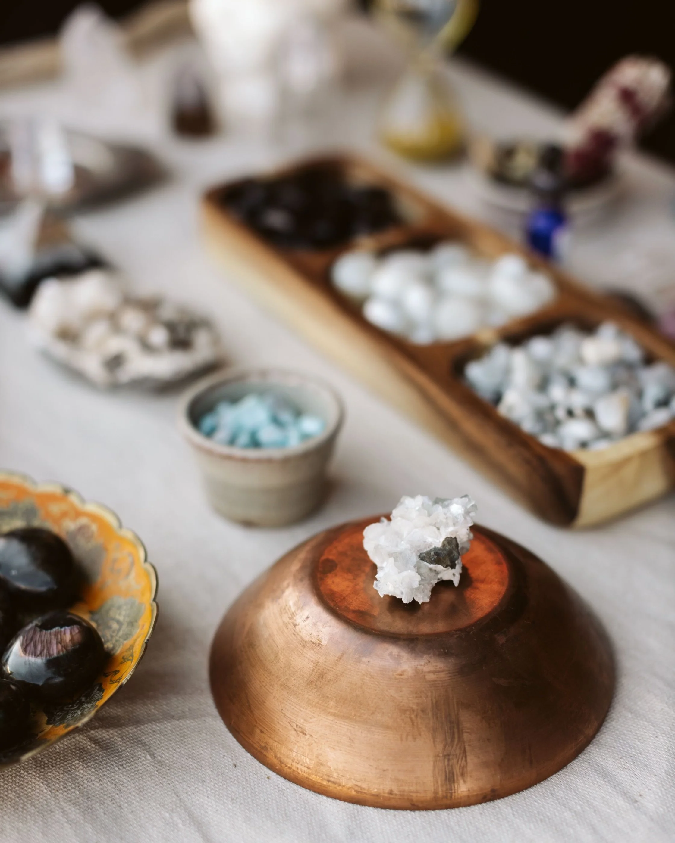 A collection of mineral and crystal specimens on a table, including a small white crystal on a copper bell-shaped object, a bowl of blue-green crystals, and various mineral stones and geodes in the background.