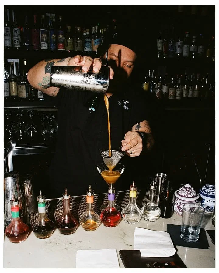 Bar with a neon sign that says "Vuze Ginten" in blue and pink, brick walls, multiple beer tap handles, bottles of liquor on shelves, and a row of bar stools.