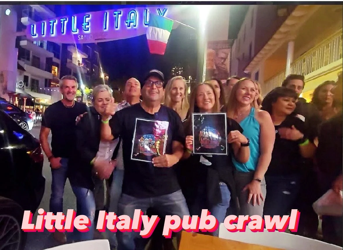 Group of people smiling and posing in front of Little Italy pub during a pub crawl at night, with neon lights and flags overhead.