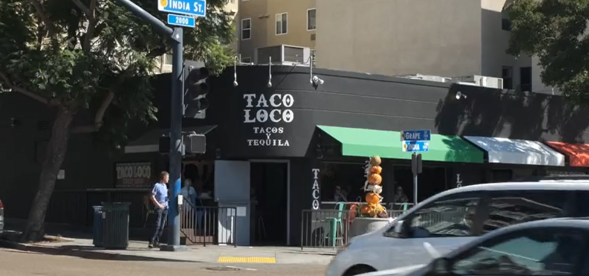 Bartender preparing cocktails at a dimly lit bar with a shelf full of liquor bottles behind him, and patrons seated at the bar.