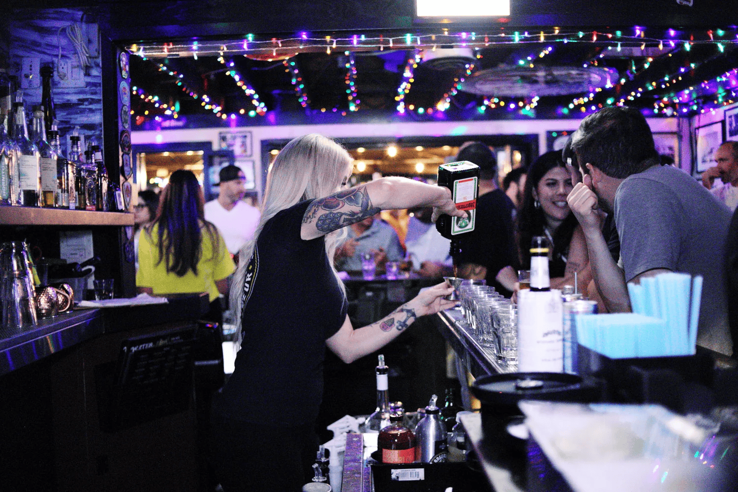 Nighttime scene outside a bar with a neon sign that reads 'Little Italy San Diego', string lights overhead, people socializing and enjoying drinks at the bar and outside.