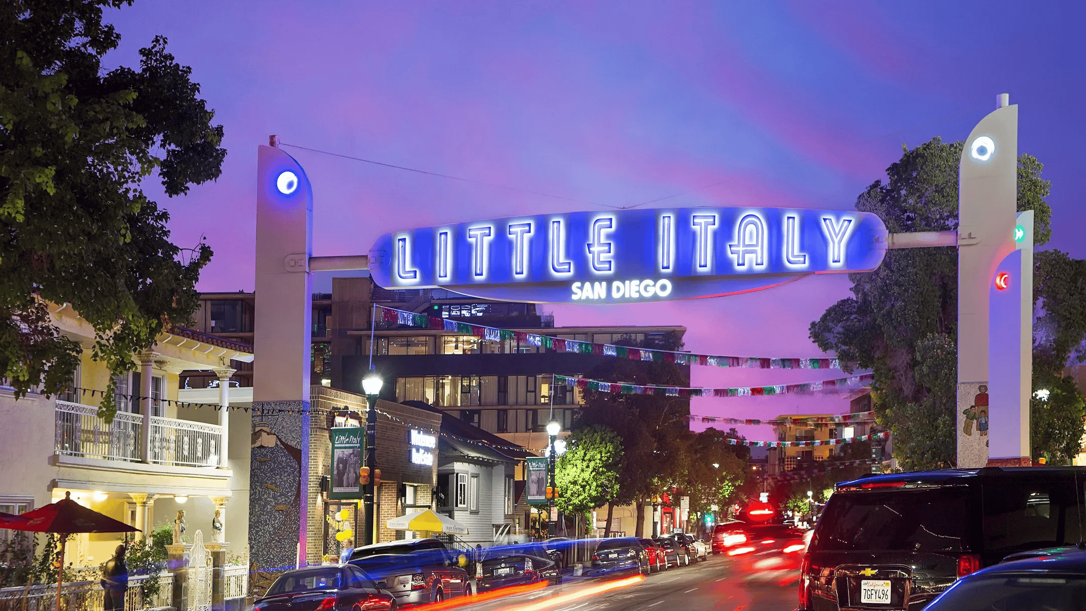 Night view of Little Italy in San Diego with bright neon sign, buildings, trees, parked cars, and streetlights against a purple and pink sky.