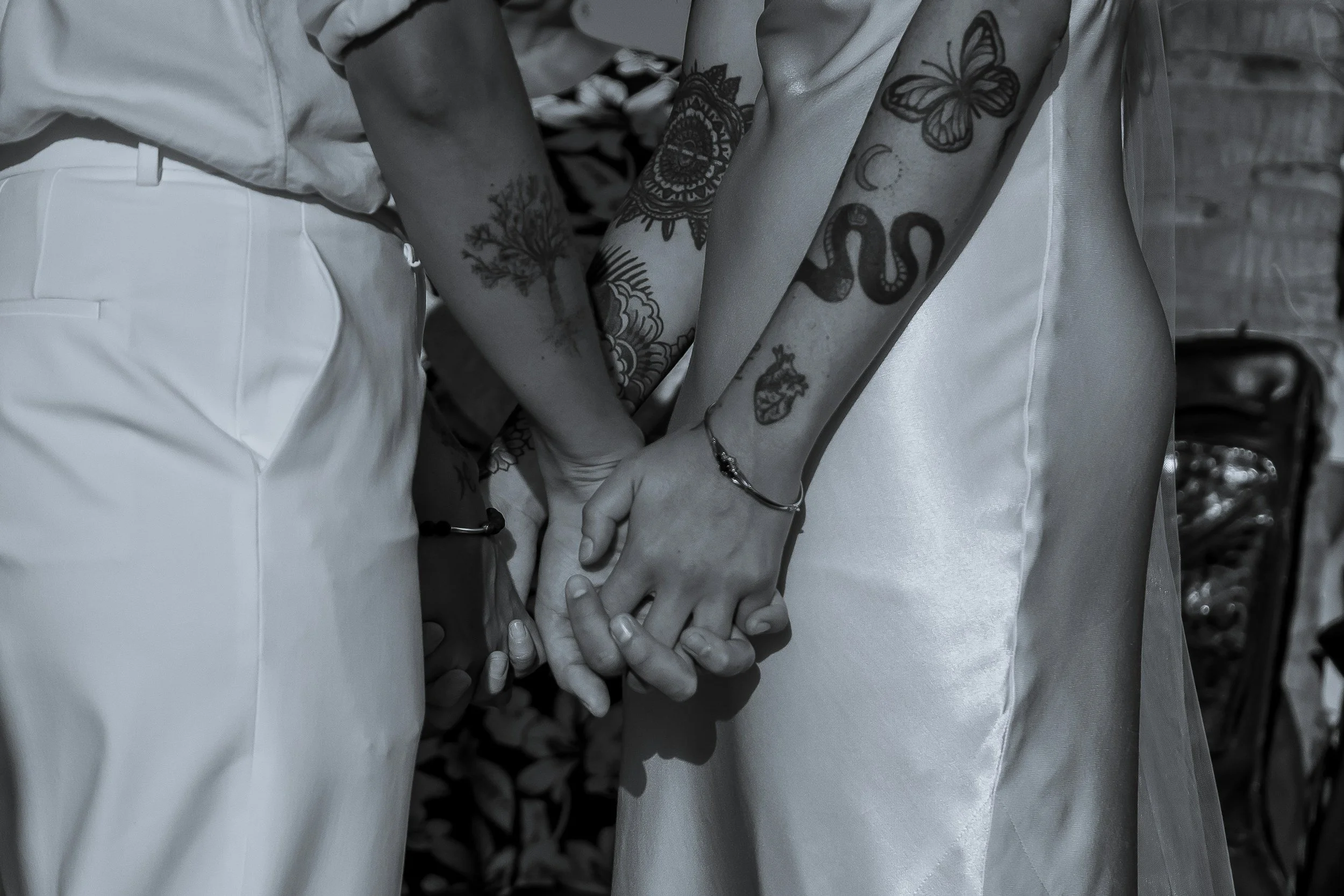 Two people holding hands, one person with tattoos on the arm and wearing a bracelet, the other person in light-colored clothing, possibly wedding attire.