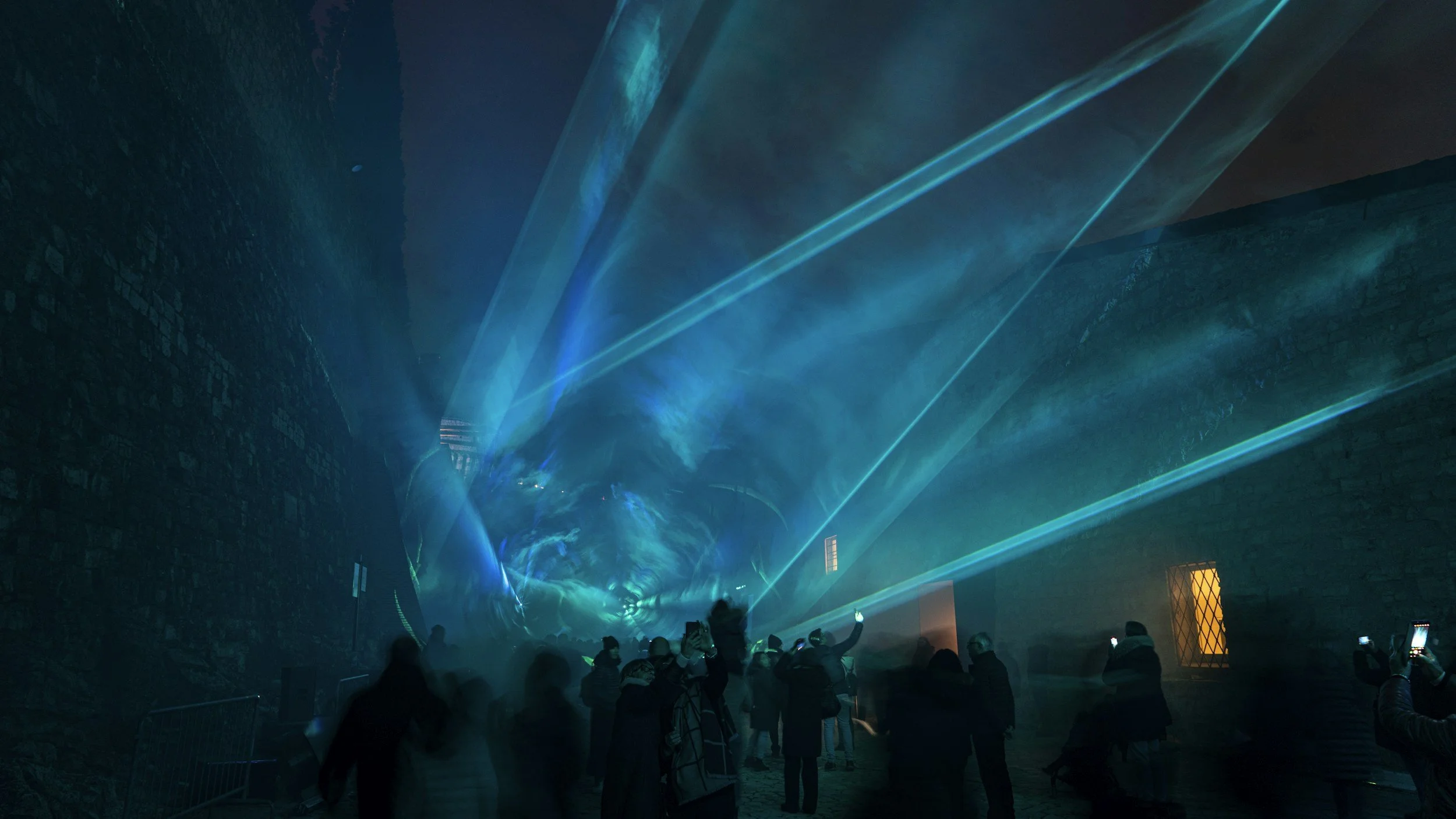 People watching a laser light show inside a dark, tunnel-like brick structure with blue and green laser beams illuminating the space.