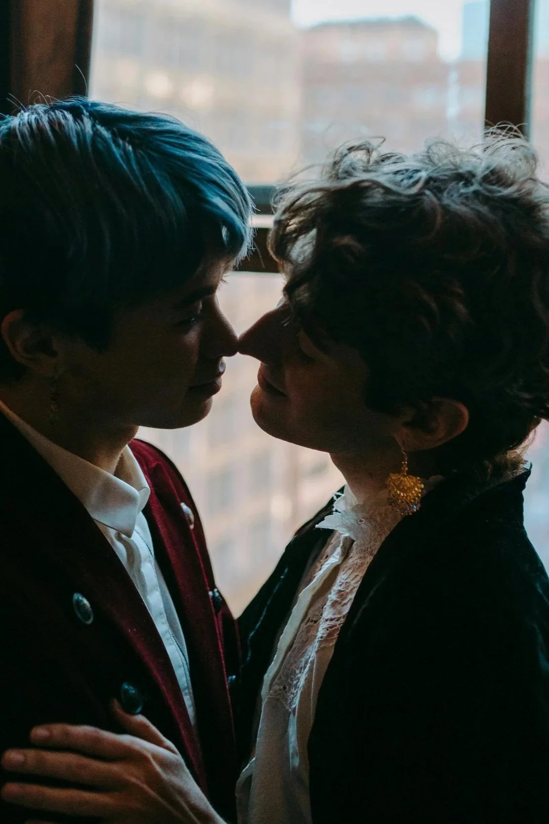 Queer couple, are close together in a dimly lit room with a window behind them. They appear to be about to kiss, with noses touching softly and eyes closed or lowered.
