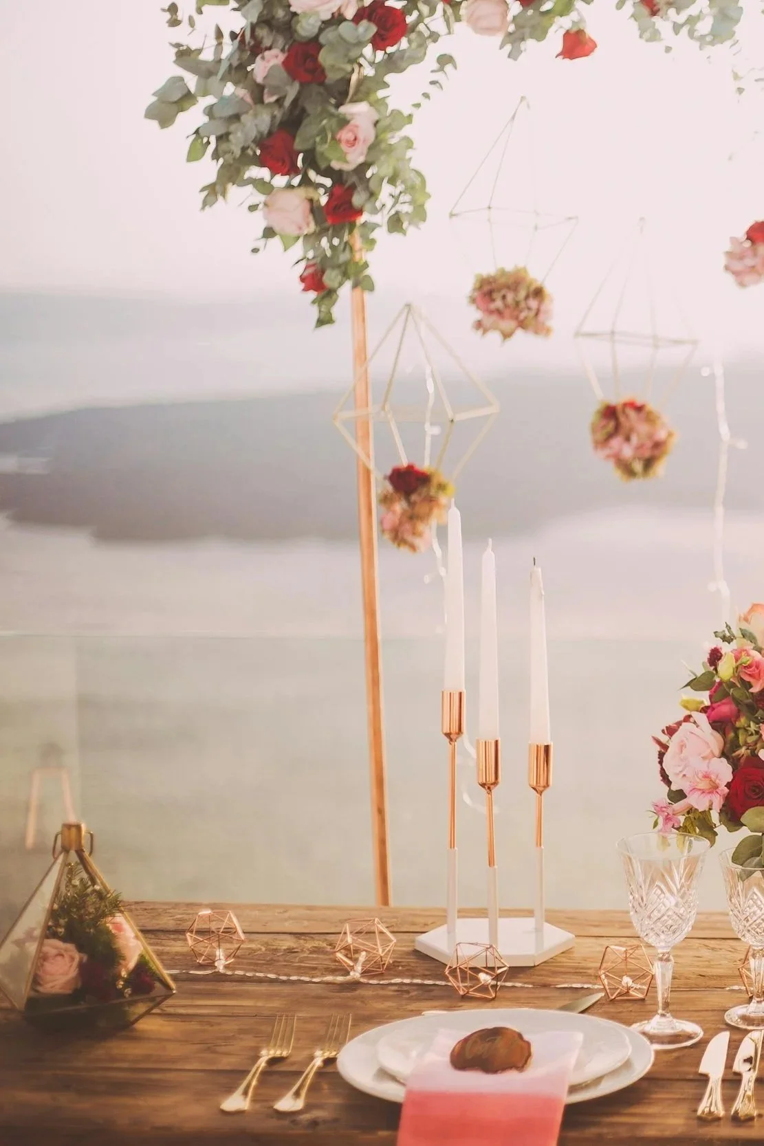 A decorated table for a special occasion with floral arrangements, tall candles, geometric ornaments, and a dessert on a plate.