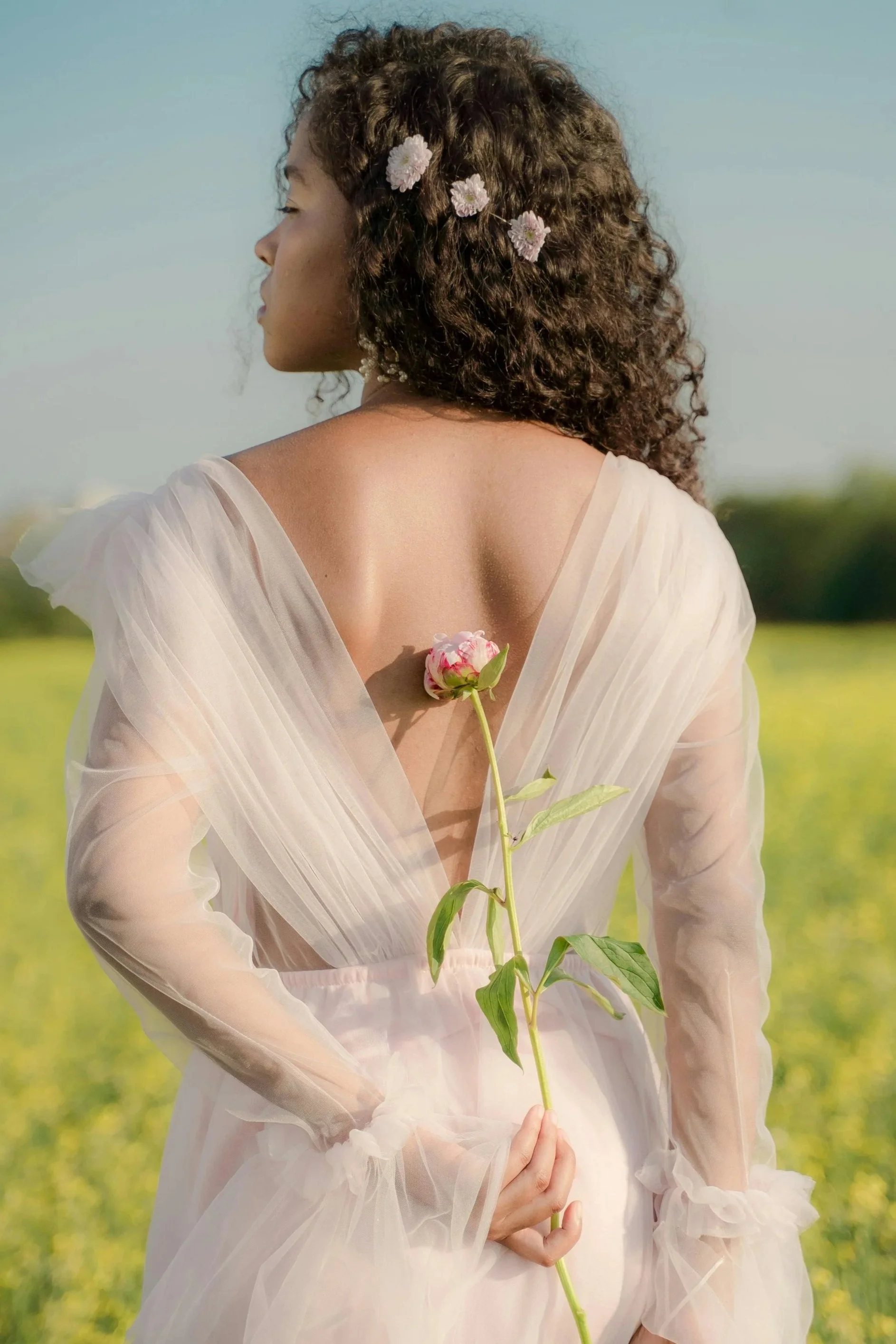 A black woman with her hair decorated with pink flowers, holding a pink flower behind her back, and wearing a sheer, flowing wedding dress outdoors.