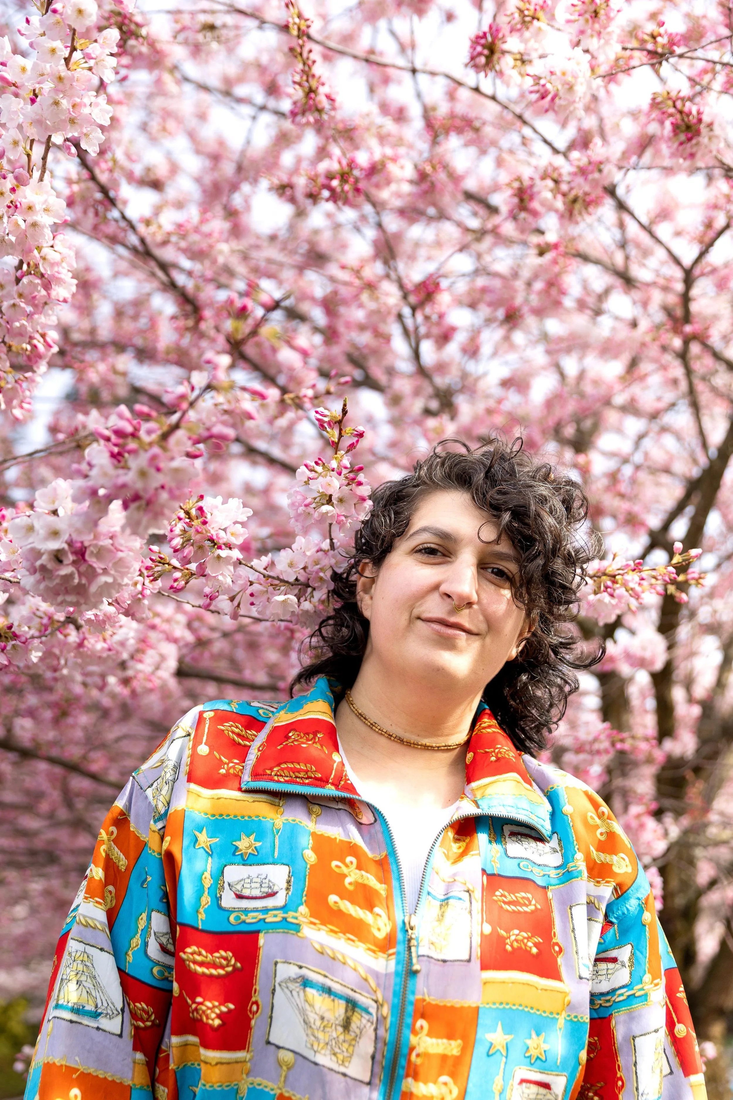 A person with short, curly dark hair and a nose ring smiling while standing outdoors among blooming pink cherry blossom trees, wearing a colorful patterned jacket.