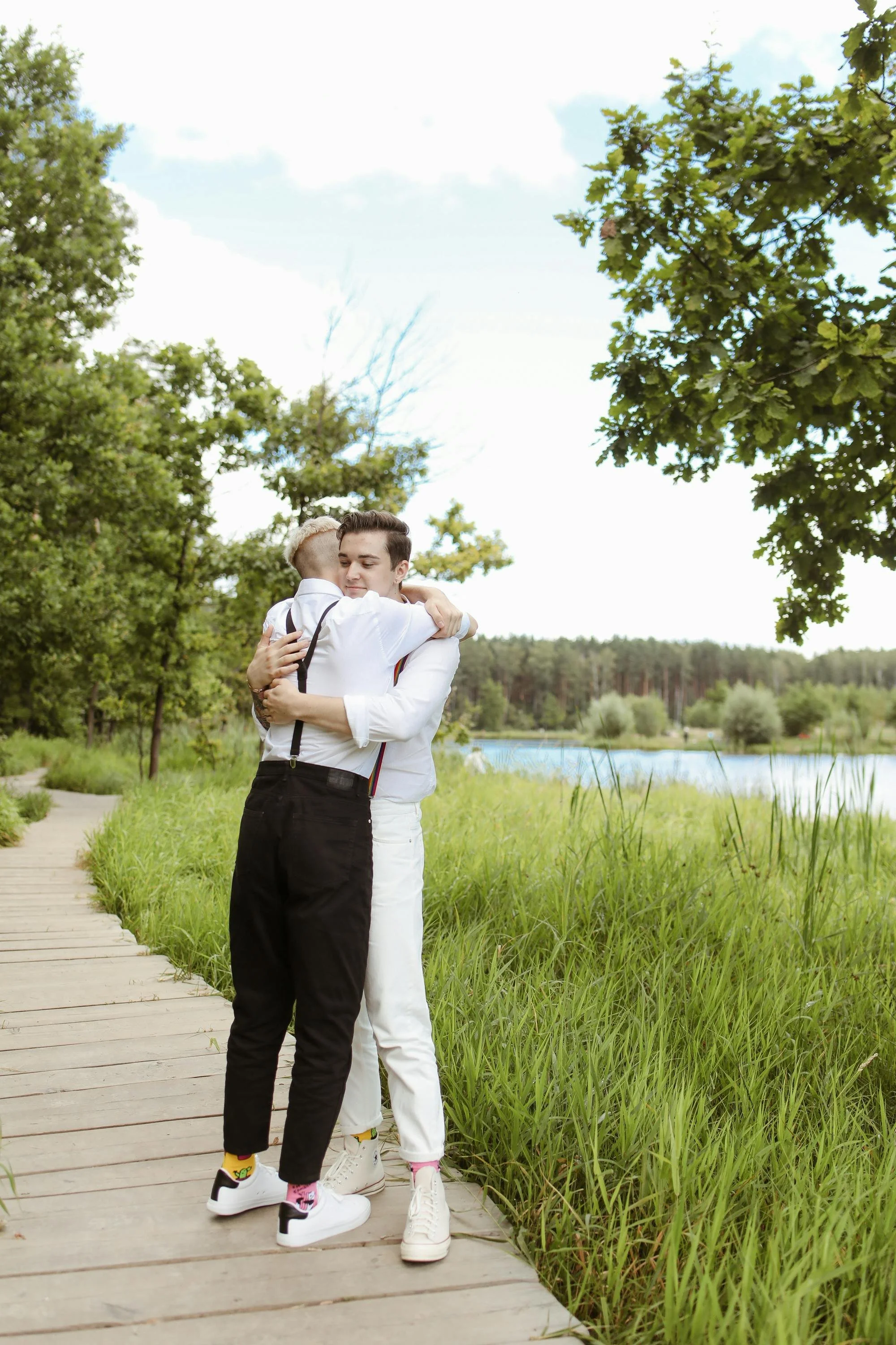 Two men hugging on a wooden walkway beside a river, surrounded by green trees and grass on a partly cloudy day.