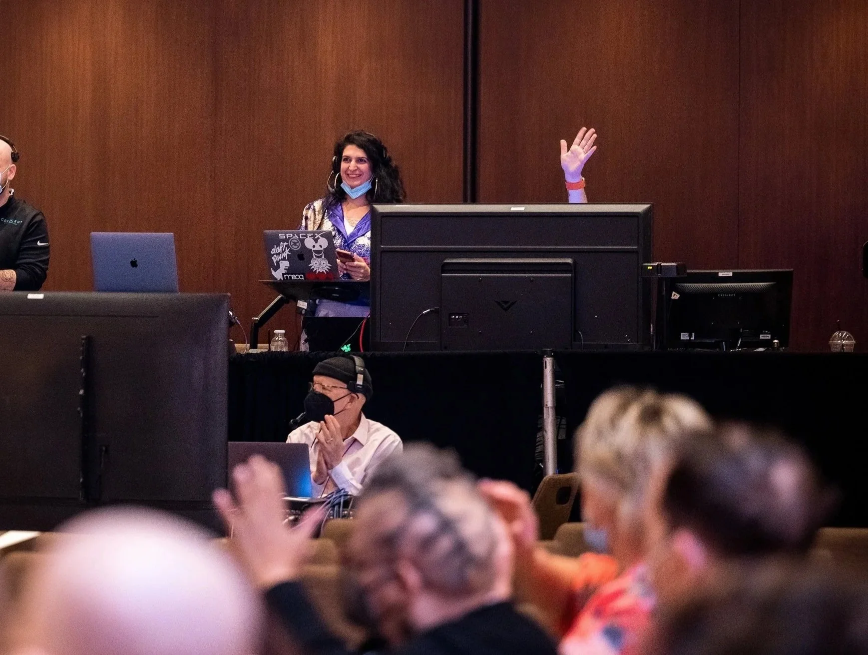 Conference room scene with a smiling and raising her hand, wearing a blue face mask around her neck, standing behind a desk with a laptop and monitors, and a man wearing a face mask and headphones seated in front of a computer. Audience in the foreground.