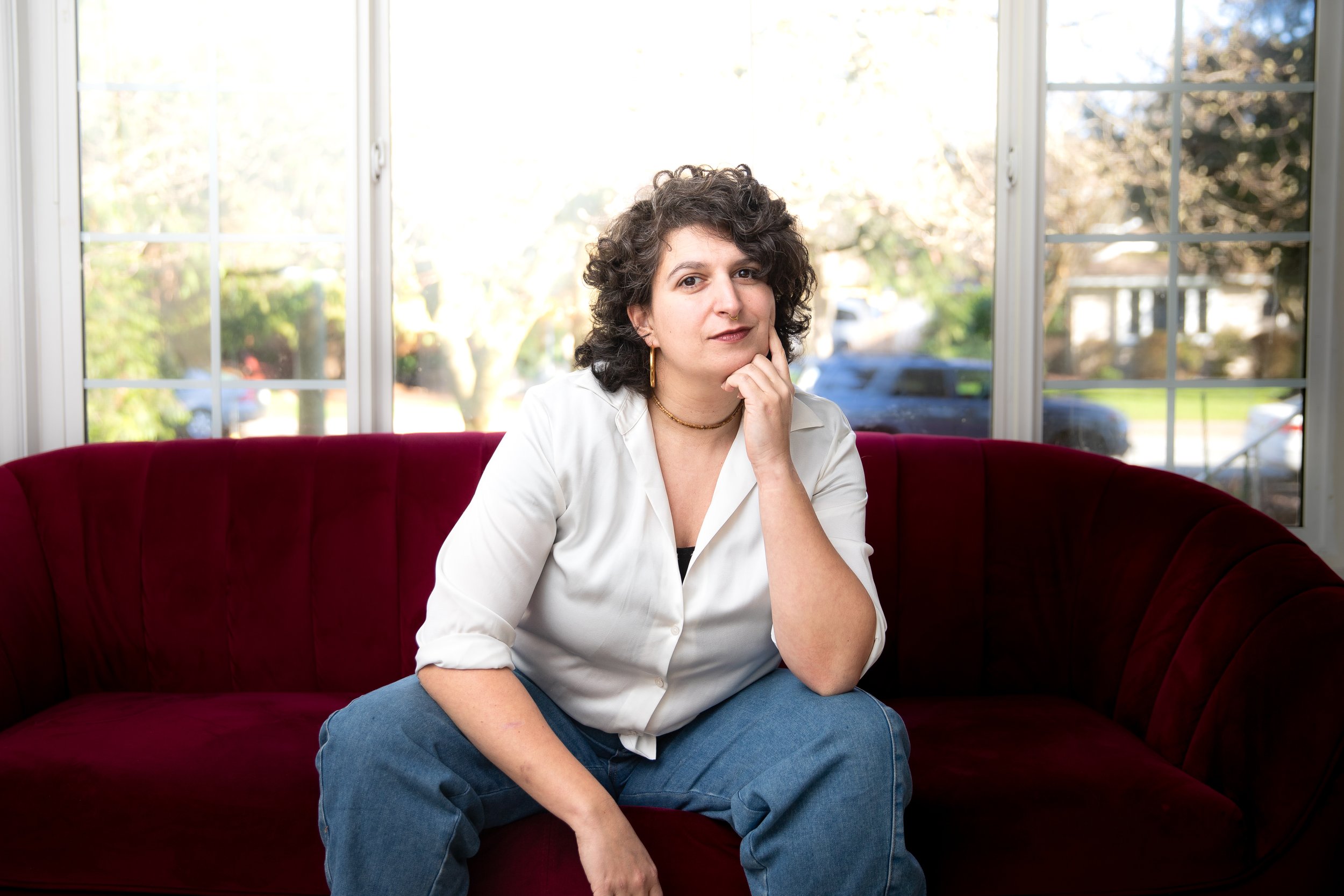 Woman with curly dark hair sitting on a red velvet couch, wearing a white shirt and blue jeans, resting her chin on her hand inside a room with large windows and a garden view.