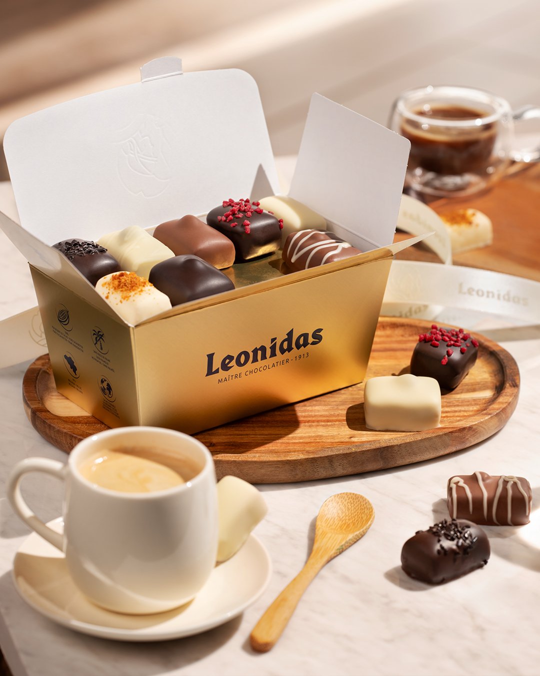 A box of assorted chocolates on a wooden tray with a cup of coffee, a small wooden spoon, and additional chocolates in front.