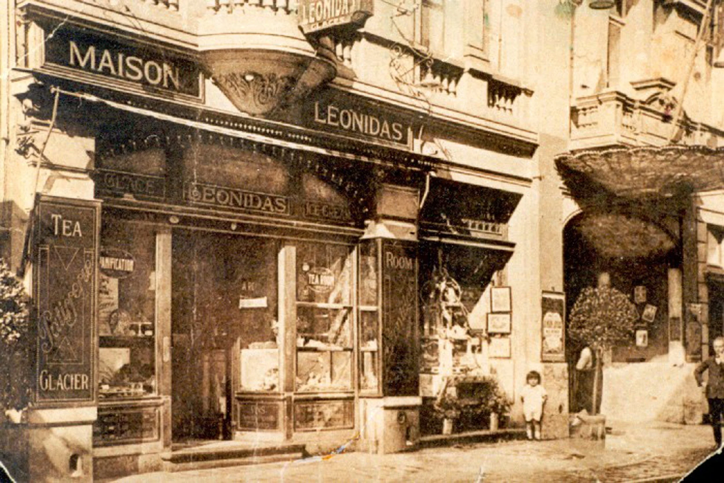 An old sepia-tone photograph of a storefront called "LEONIDAS" with signage indicating it sells tea, glaces, and other goods. There are a few people, including a small child, standing outside on the sidewalk in front of the store.