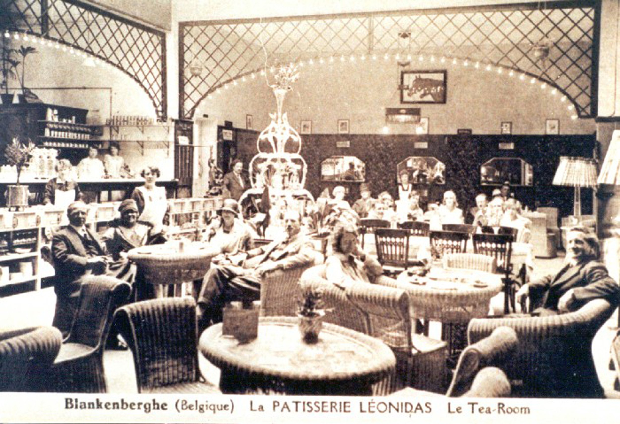 Black and white vintage photograph of the tea room at La Patisserie Léonidas in Blankenberghe, Belgium. The room is filled with people sitting at tables, with a multiple-tiered fountain in the center. The decor includes arched windows, wall-mounted shelves, and framed pictures on the walls.