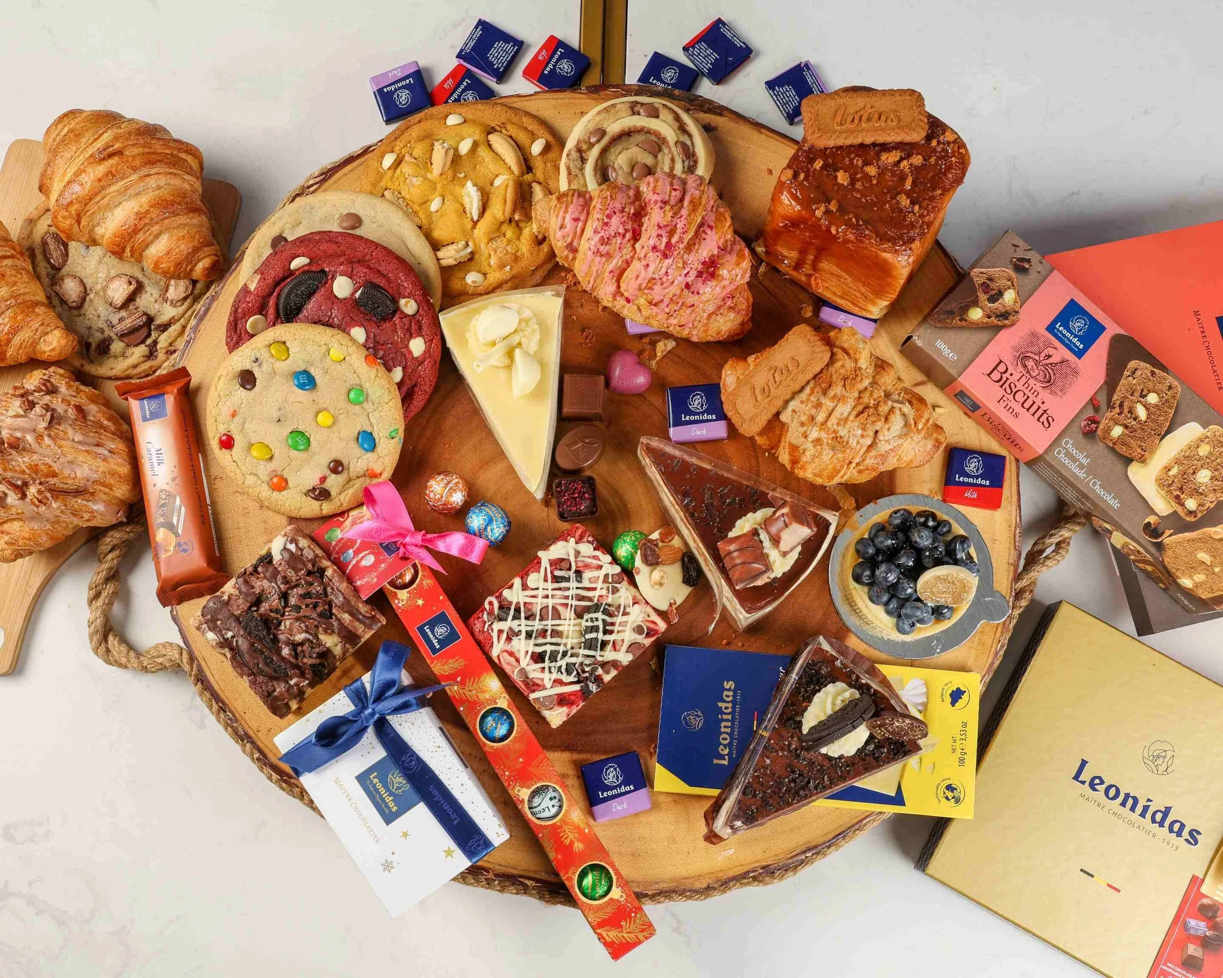 A round wooden tray filled with assorted cookies, chocolates, and pastries, including croissants, layered cake slices, and chocolate bars, along with various boxed chocolates and candy.