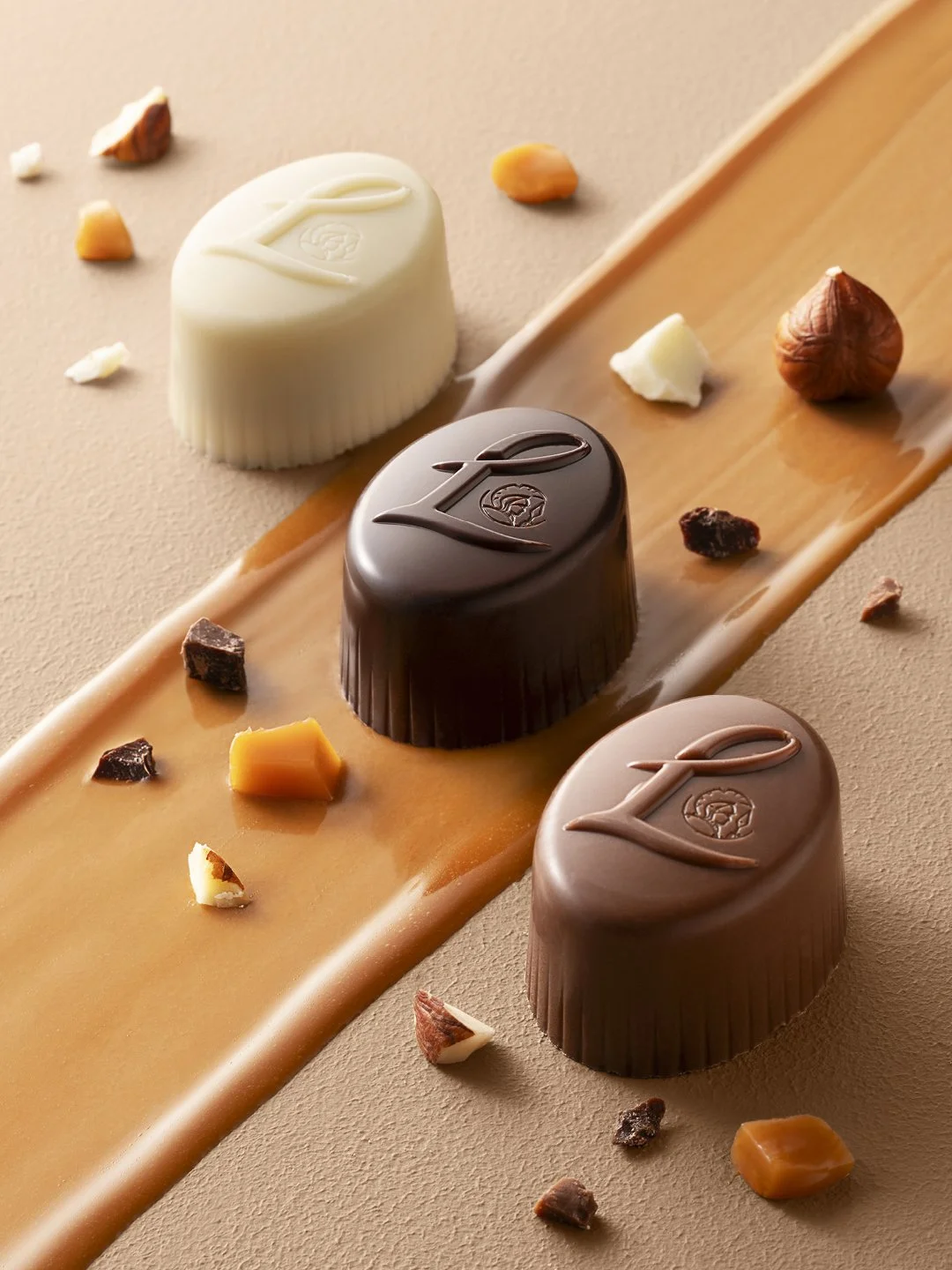 Three chocolates with an "L" on top, arranged on a caramel and white chocolate drizzle, surrounded by assorted nuts and small chocolate pieces.