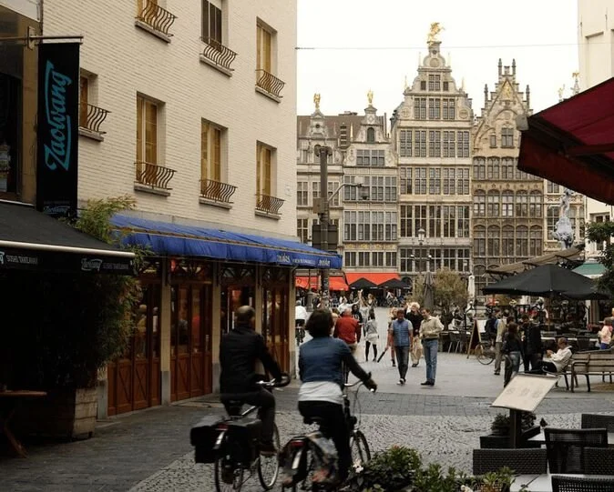 People biking and walking in a lively European city square with historic buildings and outdoor cafes.