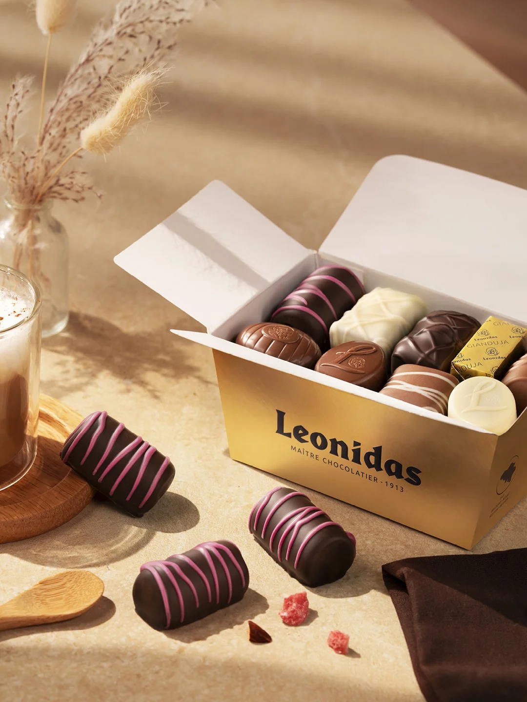 Box of assorted chocolates on a table with a few chocolates outside the box, a wooden spoon, a glass of drink, and a vase with dried plants.