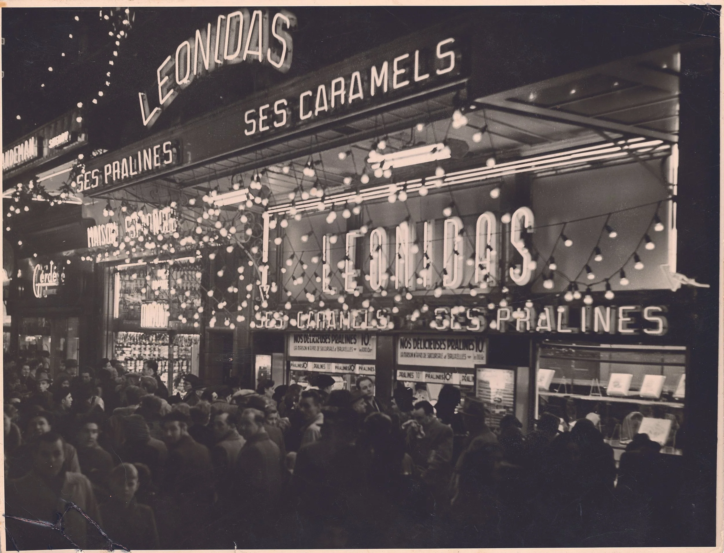 A busy street market stall with neon signs and string lights, selling pralines, caramel, and marzipan, with a crowd of people in front.