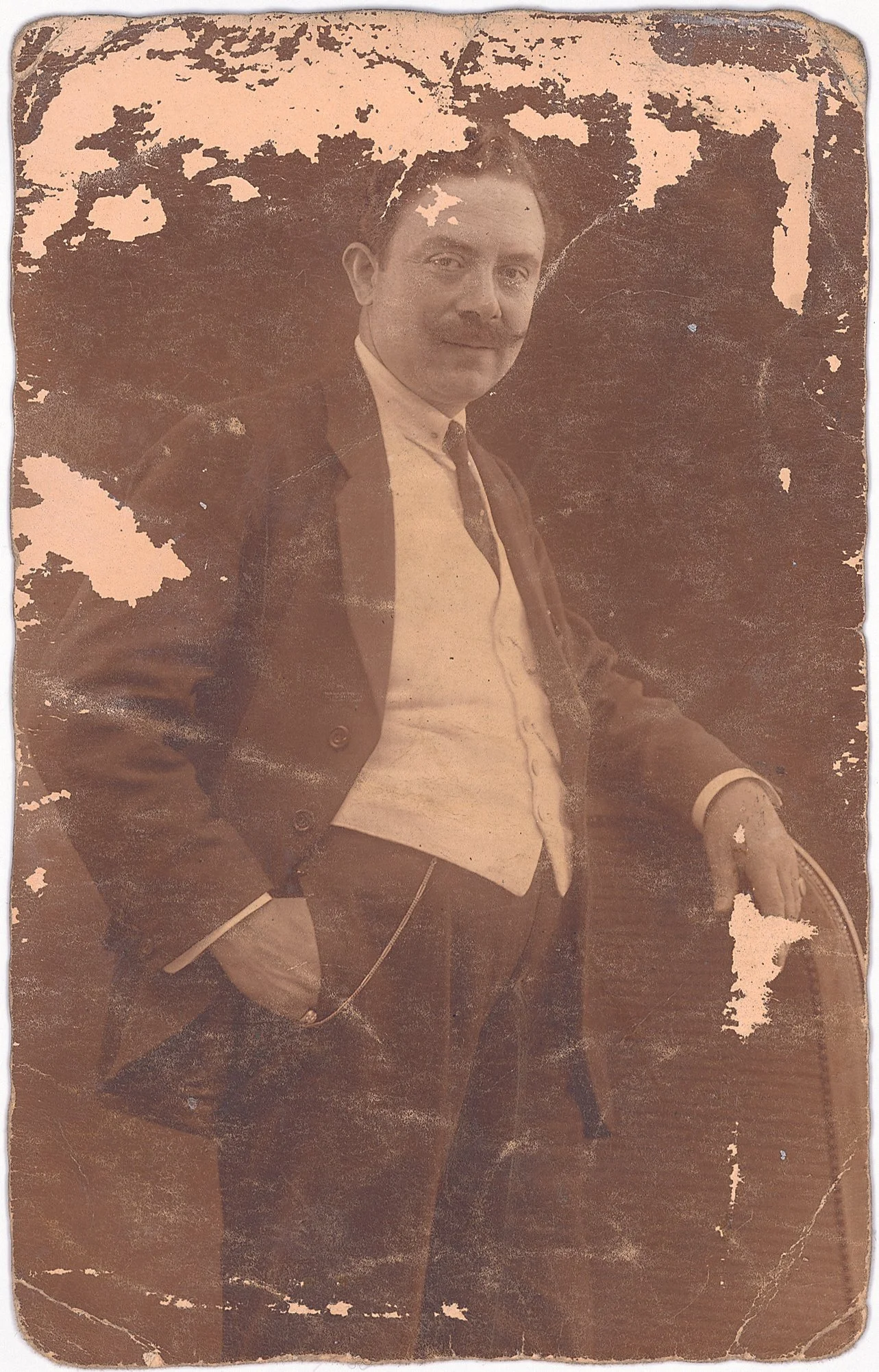 An old, sepia-toned photograph of a man in vintage attire, standing with one hand in his pocket and the other resting on a chair, with a worn and damaged surface.