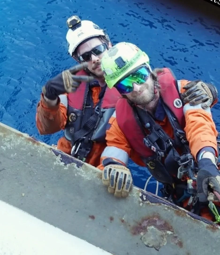 Two rescue workers in orange uniforms, helmets, and gloves are climbing above a body of water on a structure.