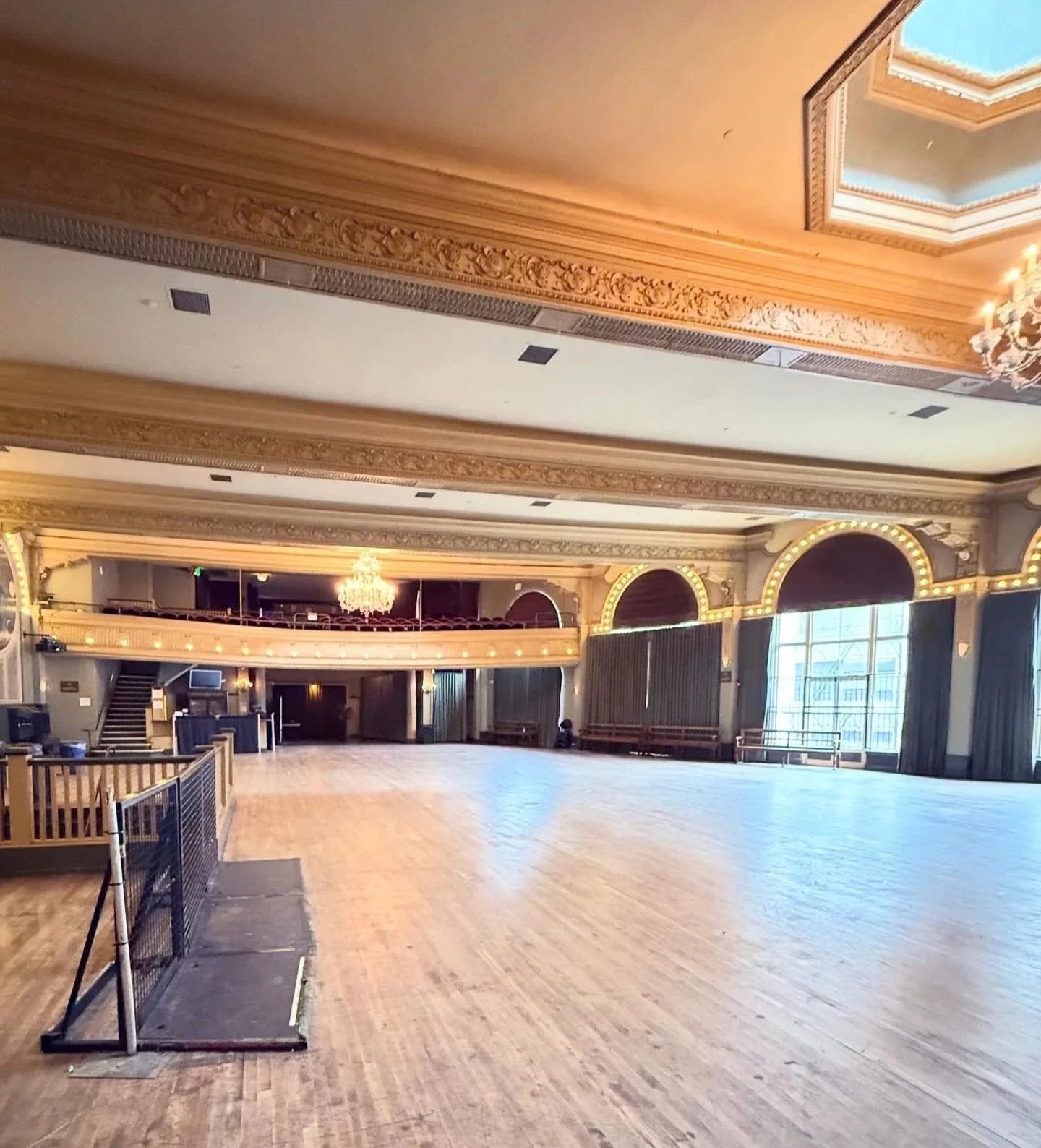 Empty ballroom with wooden floor, ornate gold ceiling, chandelier, stage area, and large windows with curtains.