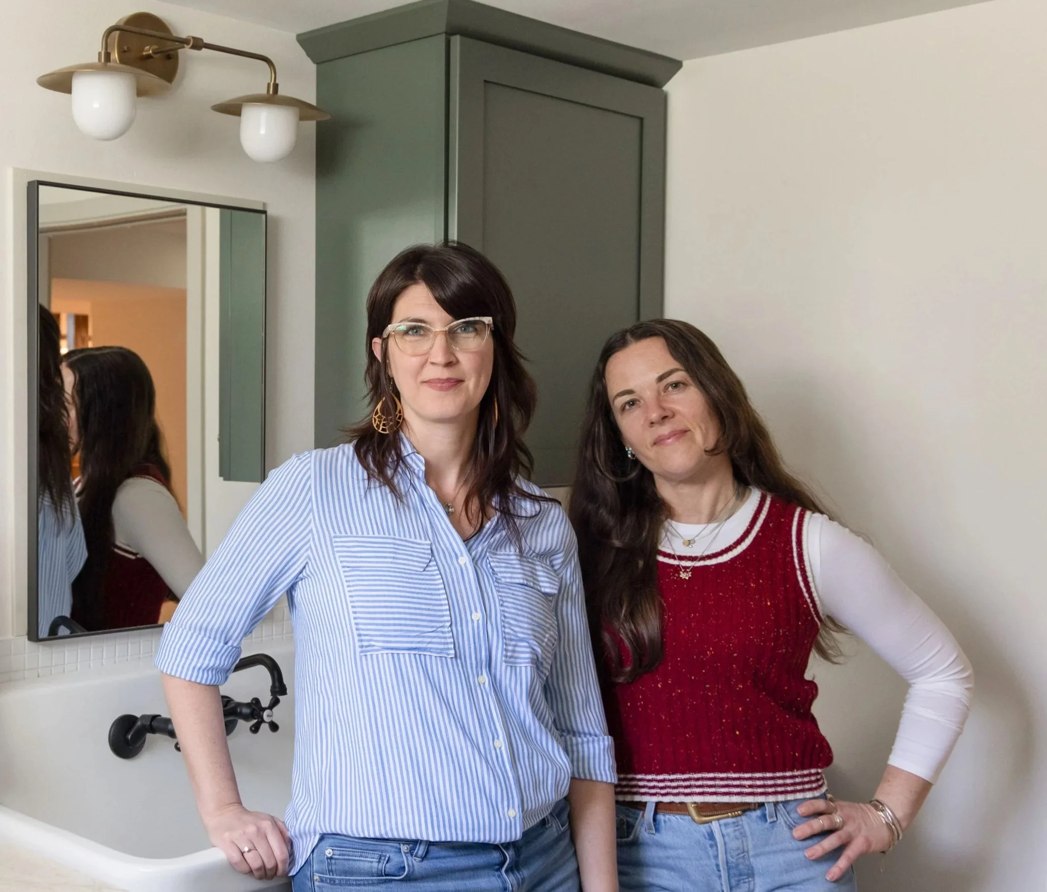 Two women standing side by side in front of a bathroom mirror, smiling at the camera. The woman on the left has shoulder-length dark hair, glasses, and is wearing a blue and white striped shirt. The woman on the right has long brown hair, and is wearing a red sweater with white stripes and a white shirt underneath. There is a green cabinet and a white wall in the background.