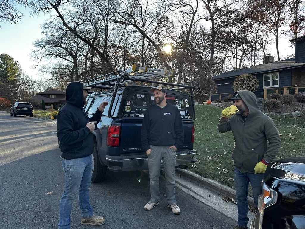 Three men are standing outside on a suburban street, engaged in conversation. They are dressed in casual hoodies and gloves, with one wearing a cap. A black pickup truck with a roof rack is parked behind them. The setting appears to be in late fall or early winter, with leafless trees and autumn-colored foliage around.