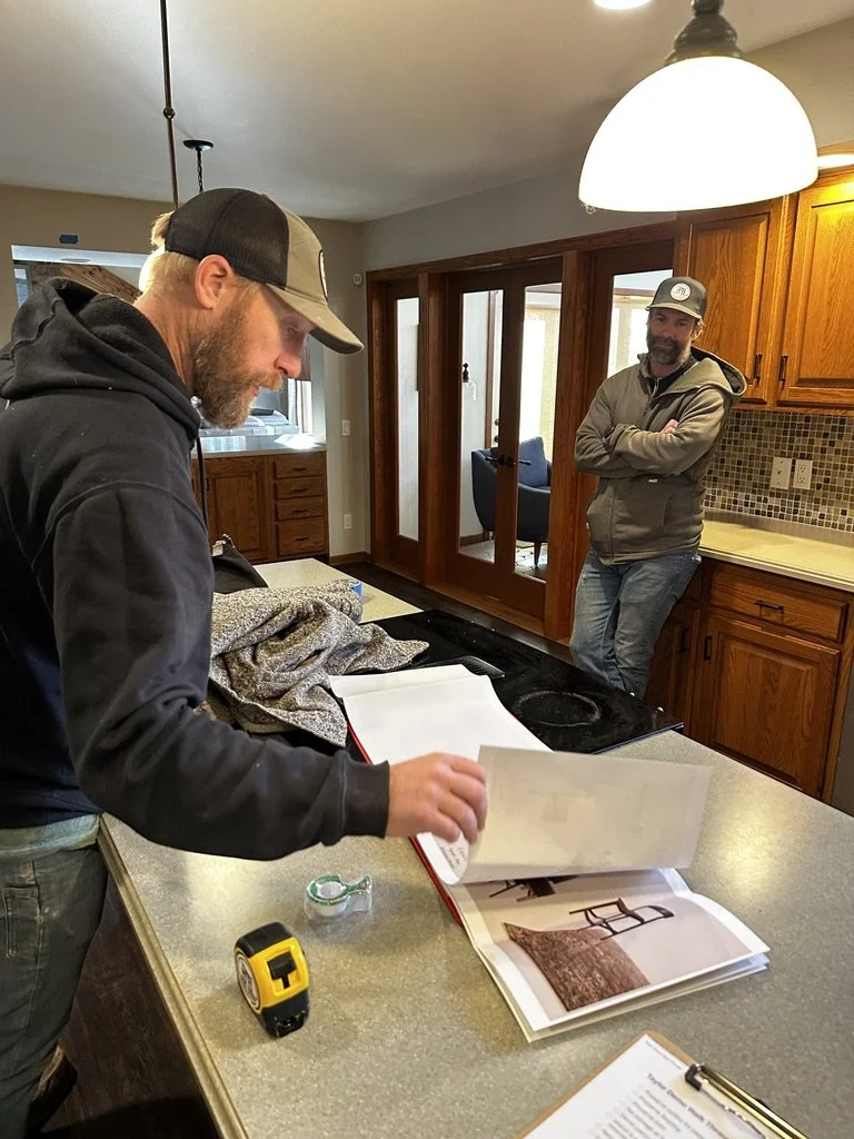 Two men in a kitchen; one is looking through a binder, while the other leans against the wooden cabinets with arms crossed.