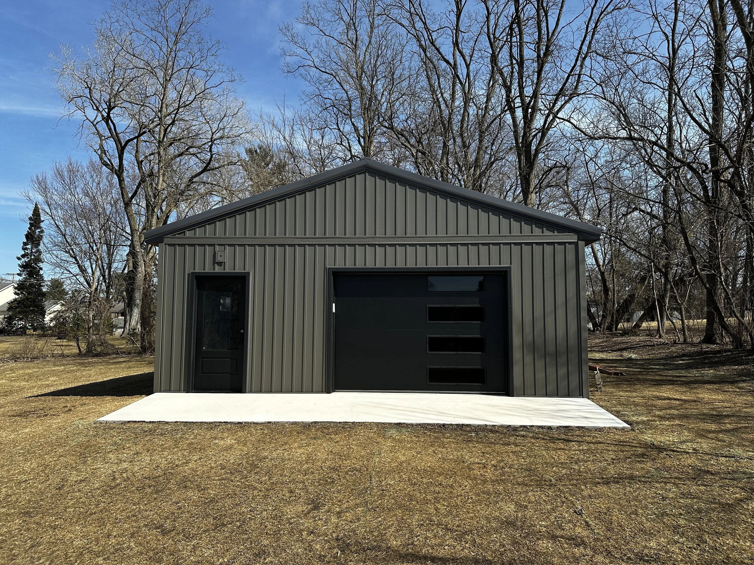 A modern gray metal shed with a flat concrete patio in front, surrounded by leafless trees under a clear blue sky.