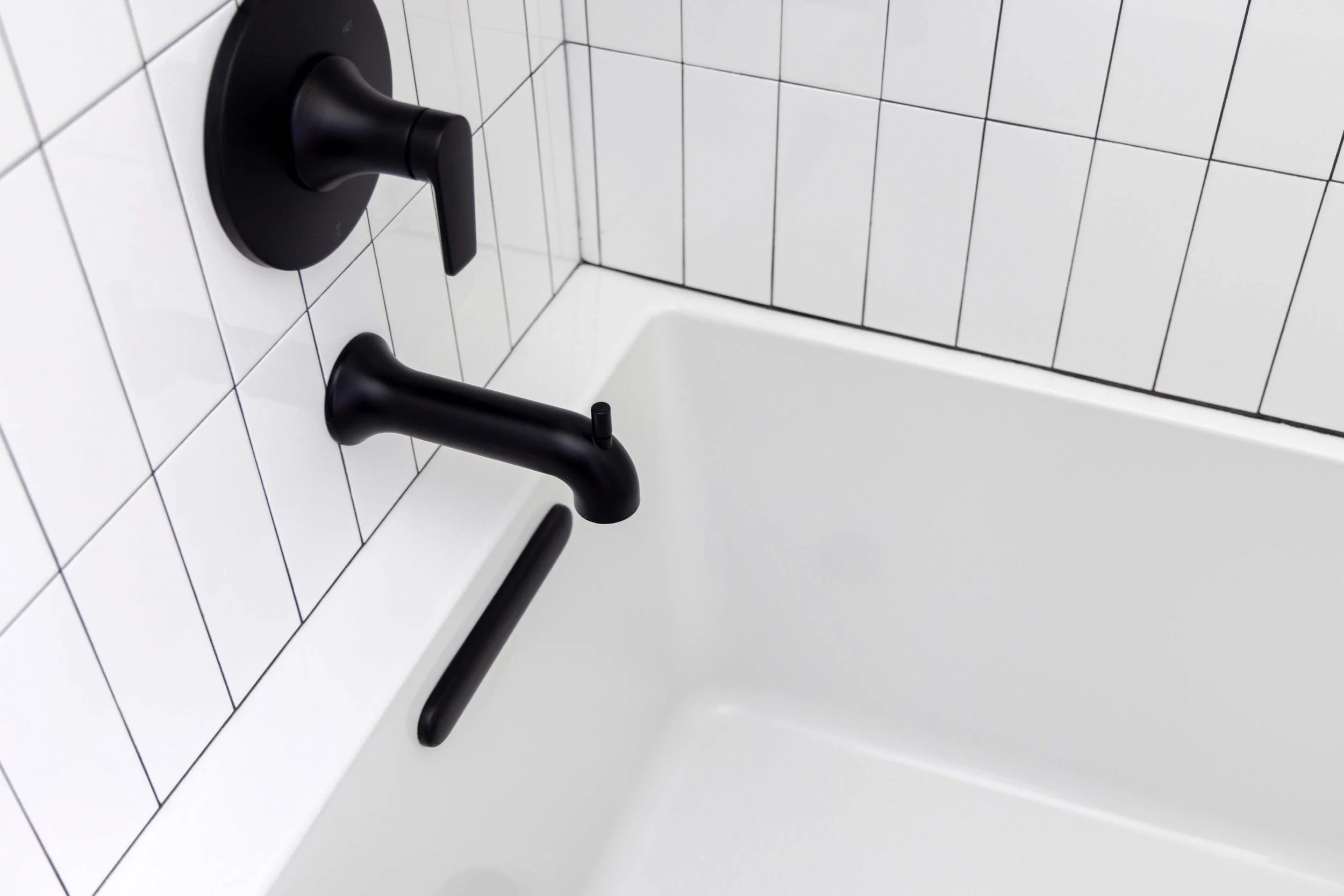 Contemporary wall-mounted matte black bathtub faucet installed against a white vertical stack tile wall.
