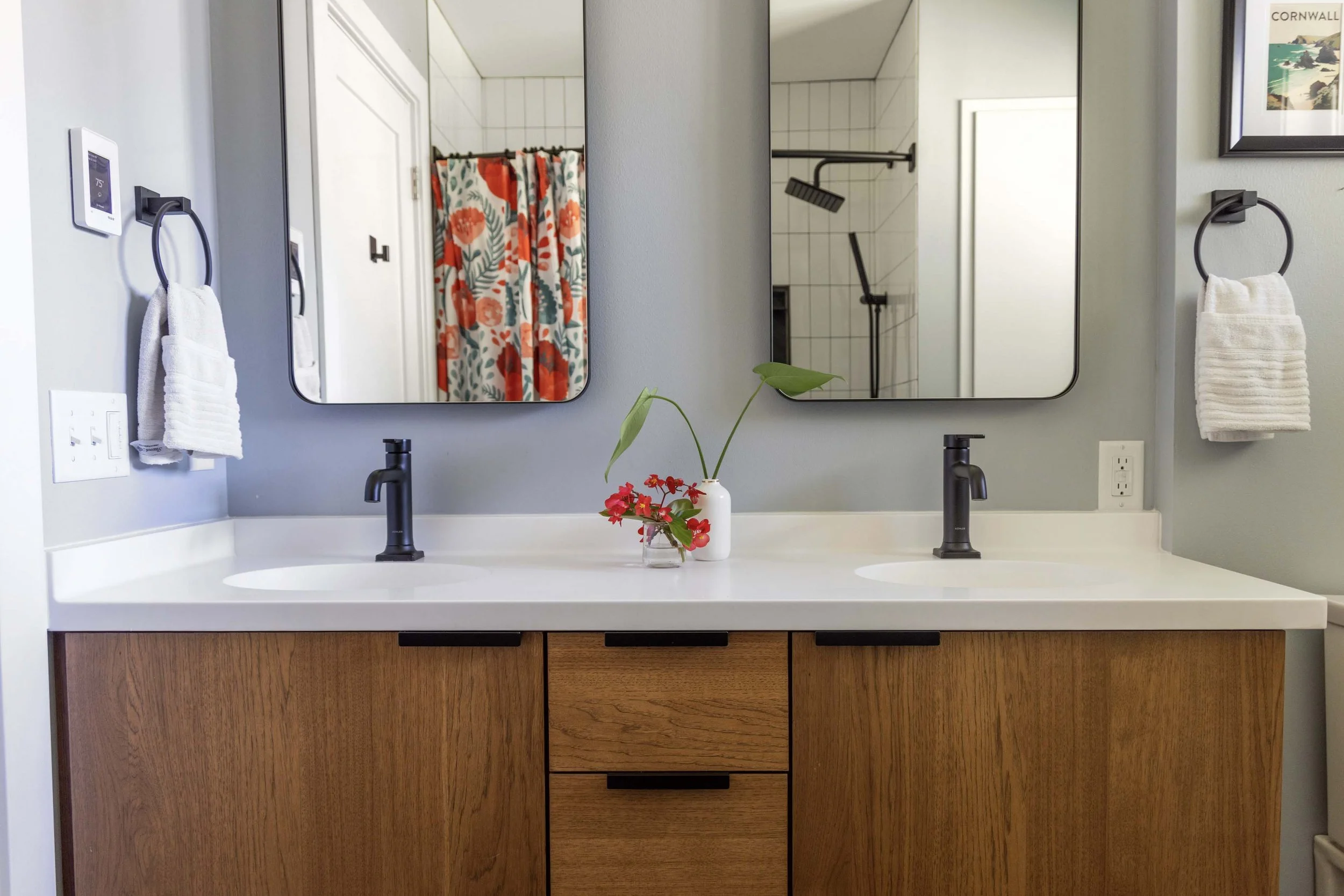 Bathroom countertop with two sinks, black faucets, a small vase with a green leaf and red flowers, large mirrors, white walls with mounted towel rings holding white towels, and a shower area with a floral curtain visible in the reflection.