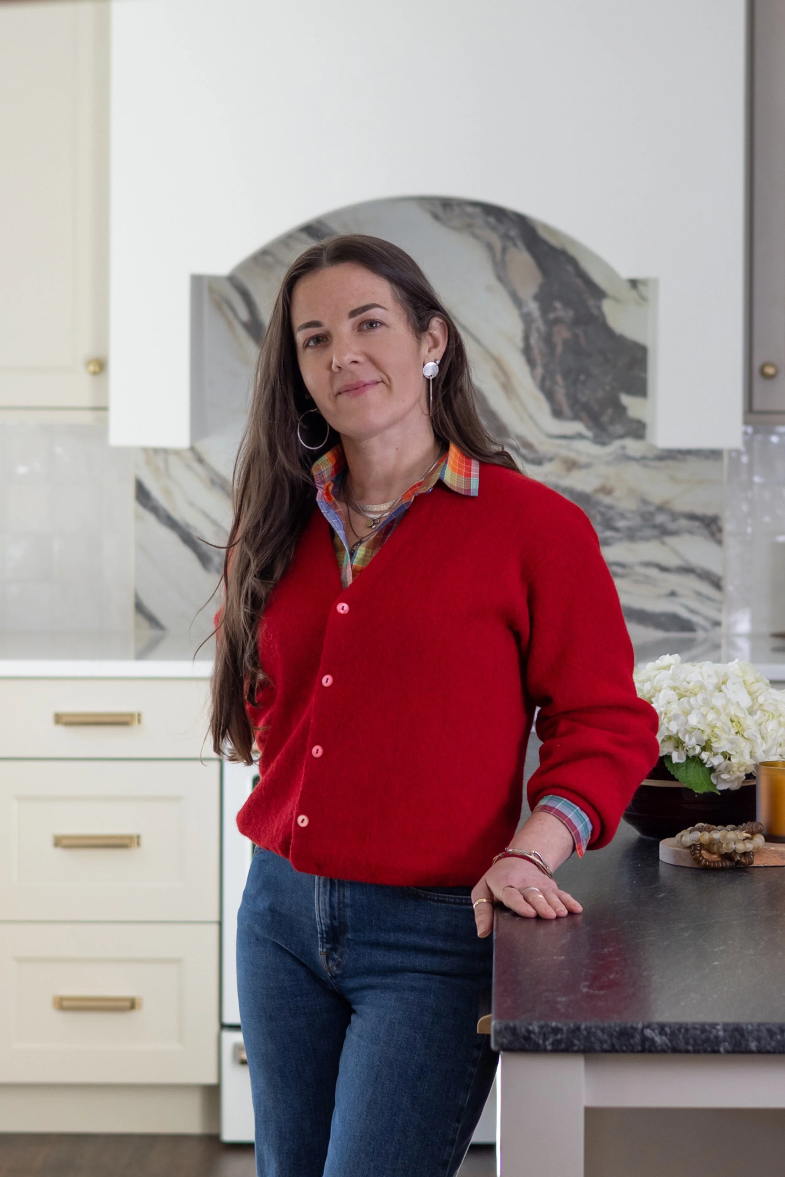 A woman with long brown hair wearing a red cardigan, plaid shirt, and jeans, standing in a modern kitchen with white cabinetry and a marble backsplash, resting her hand on the kitchen counter.