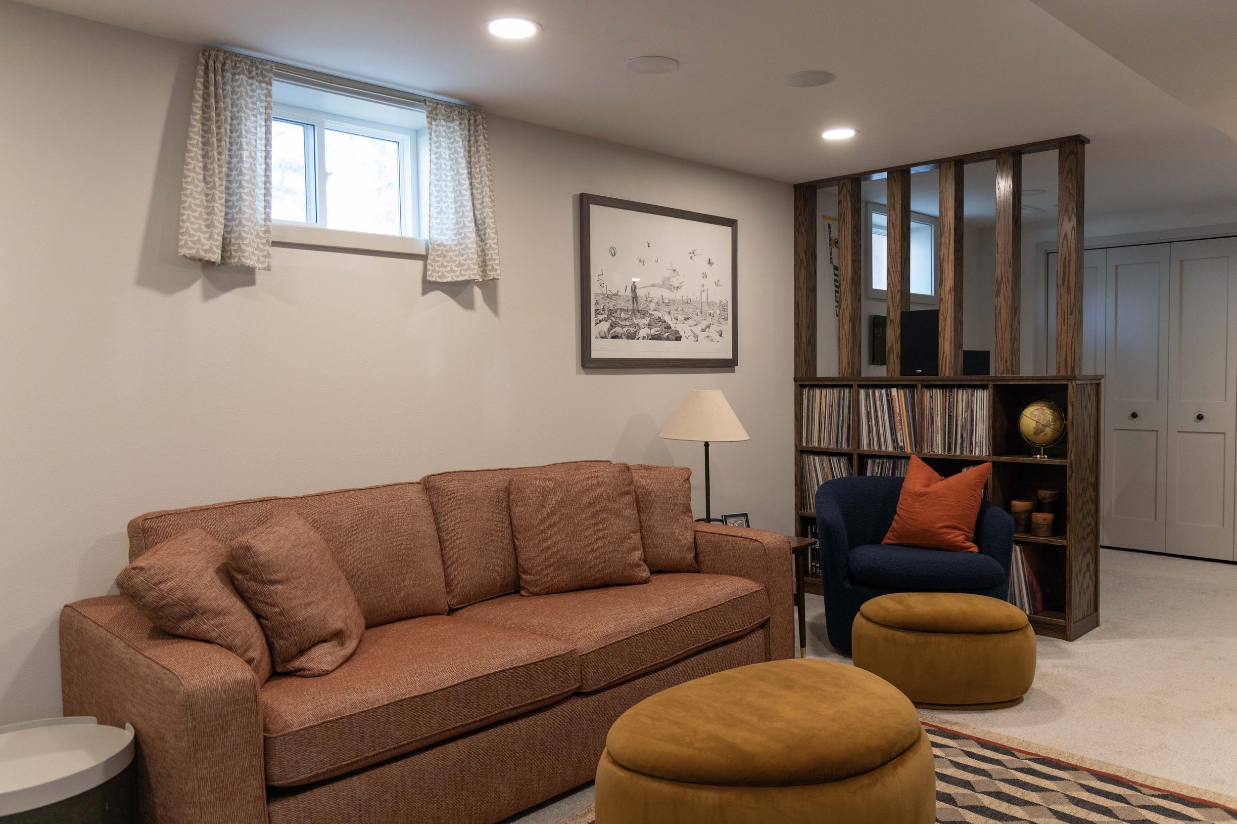 Basement living space with a terracotta sofa, round storage ottoman, and a custom slatted wood privacy screen.
