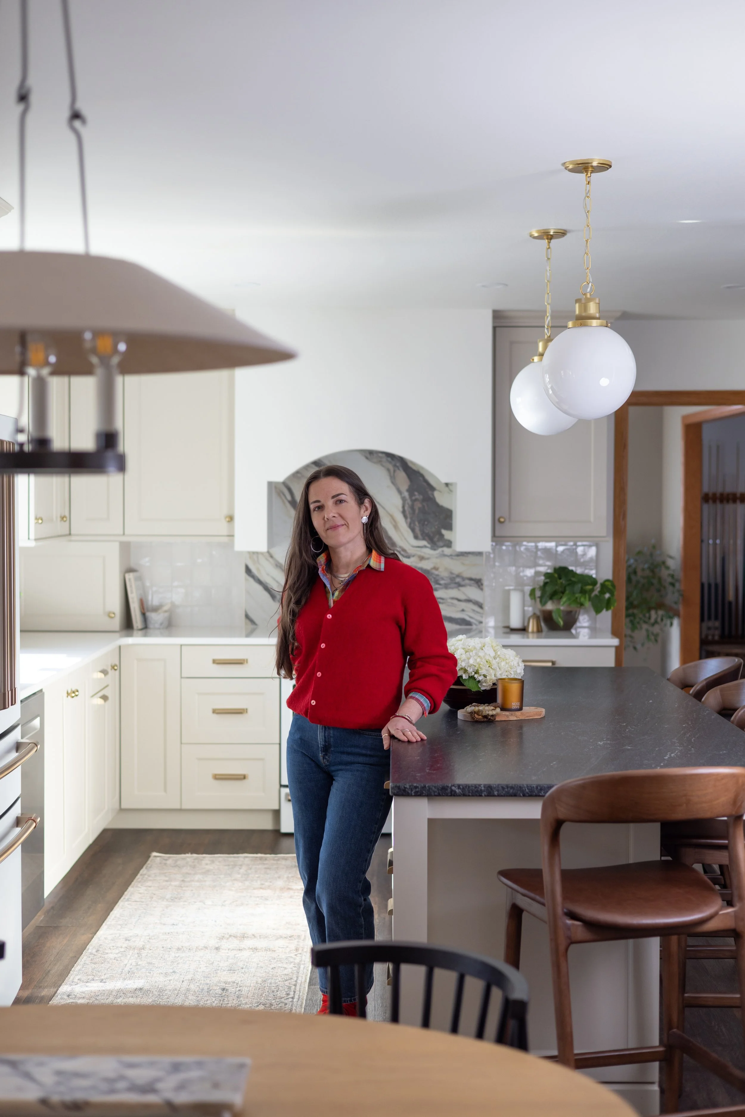 A woman with long brown hair wearing a red sweater and blue jeans standing in a modern kitchen with white cabinets, a black countertop, and a marble backsplash. She has her hand resting on the counter and looks towards the camera. The kitchen features gold and glass pendant lights, and there are plants and decorative items on the counter and shelves.