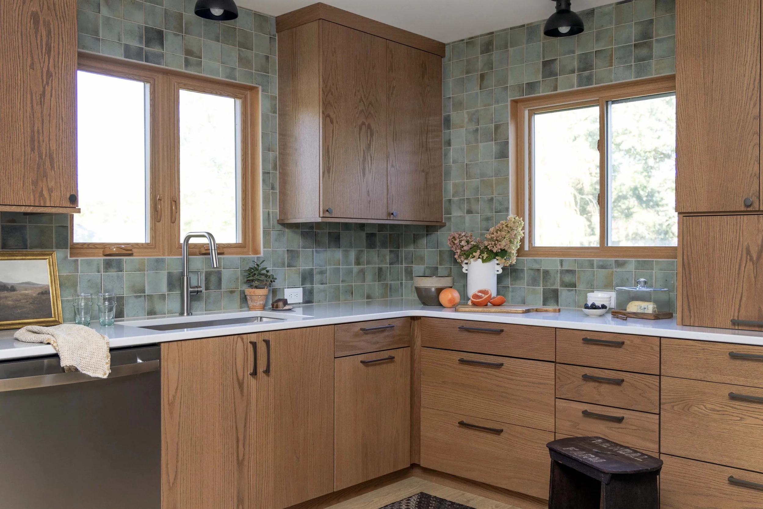 A kitchen with wooden cabinets, green tile backsplash, and two windows showing outdoor greenery. Items on the countertop include a vase with flowers, vegetables, bowls, and kitchen utensils.